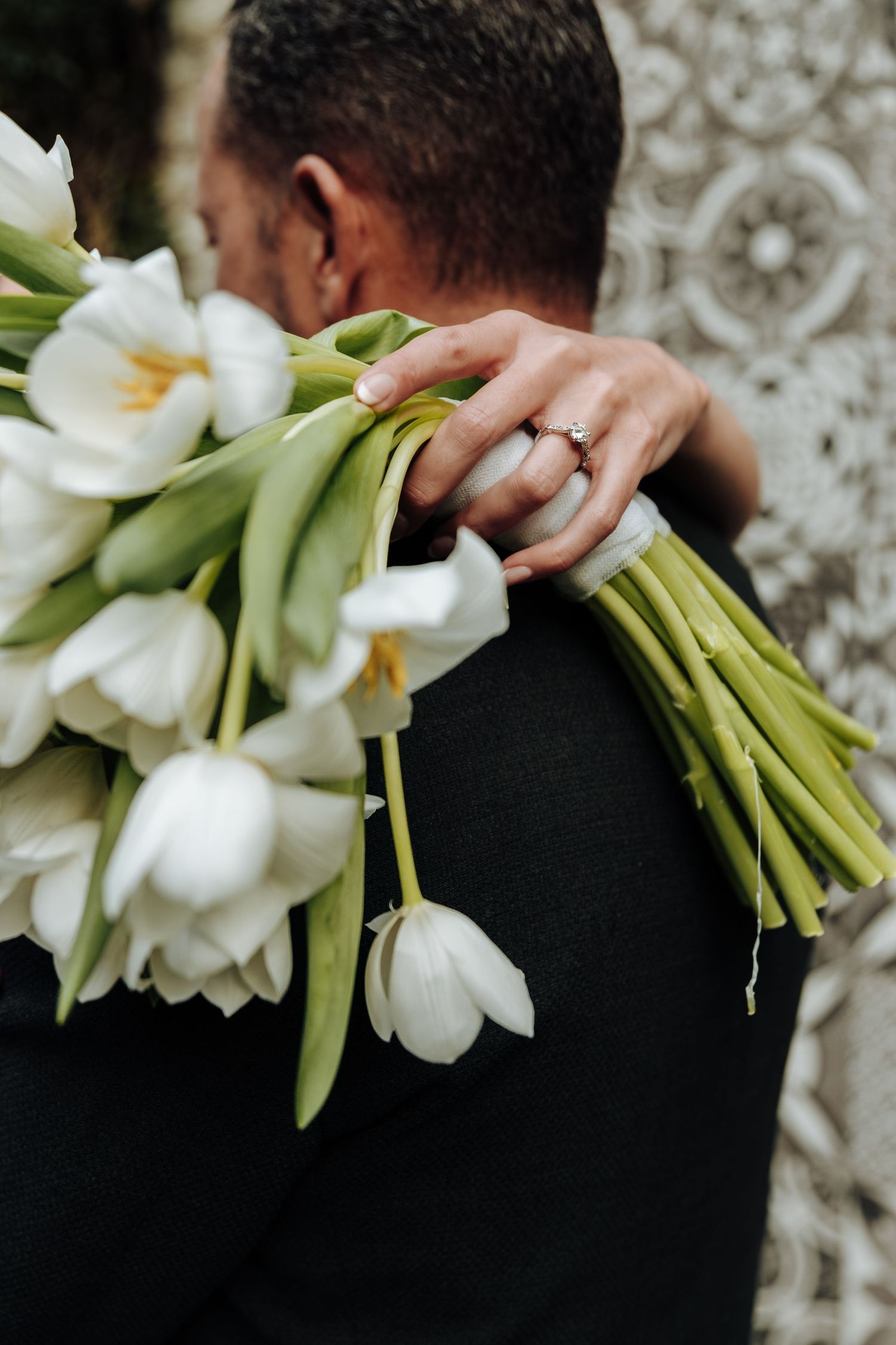 A woman wearing a diamond ring holds a bouquet of white tulips over a man's shoulder in a romantic embrace.