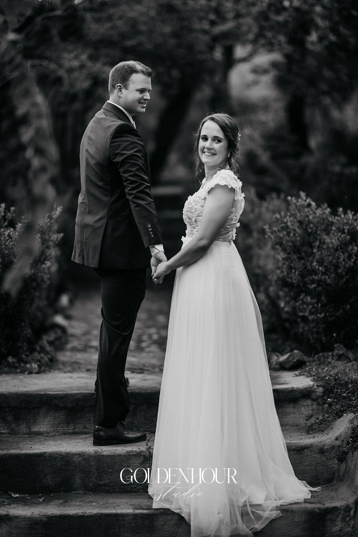 Black and white portrait of bride and groom holding hands on garden steps.