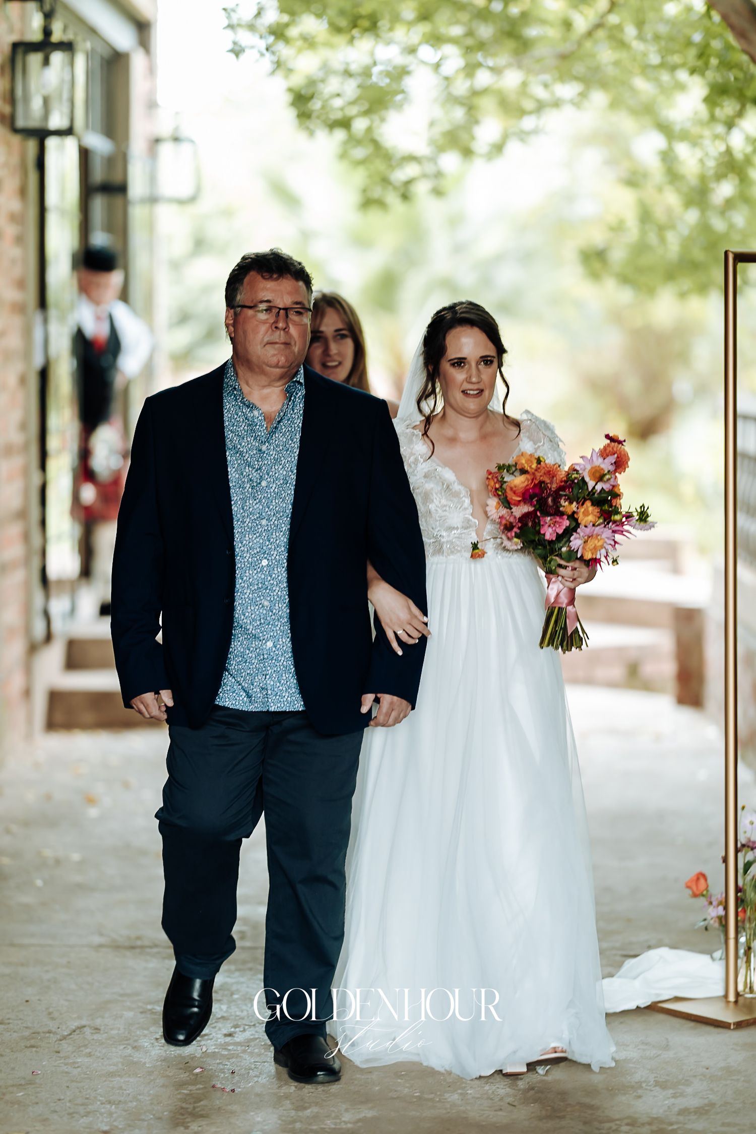 Father escorting smiling bride in lace wedding gown holding a colorful bouquet.