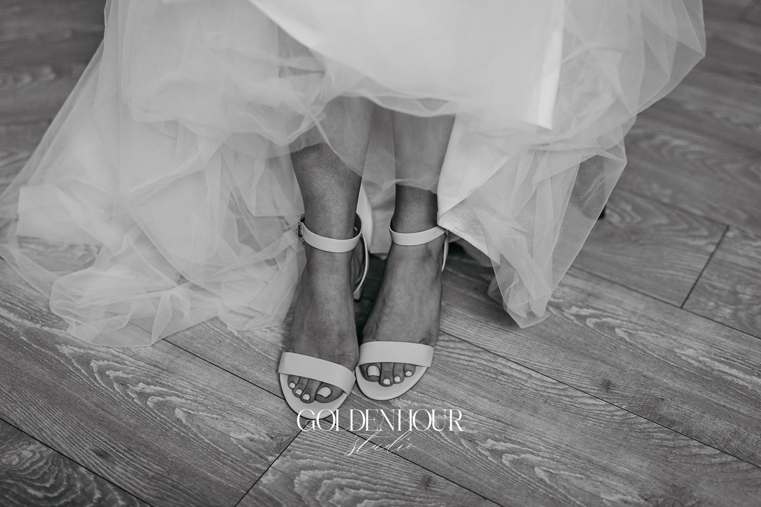 Black and white close-up of bride wearing elegant ankle-strap heels on wooden floor.