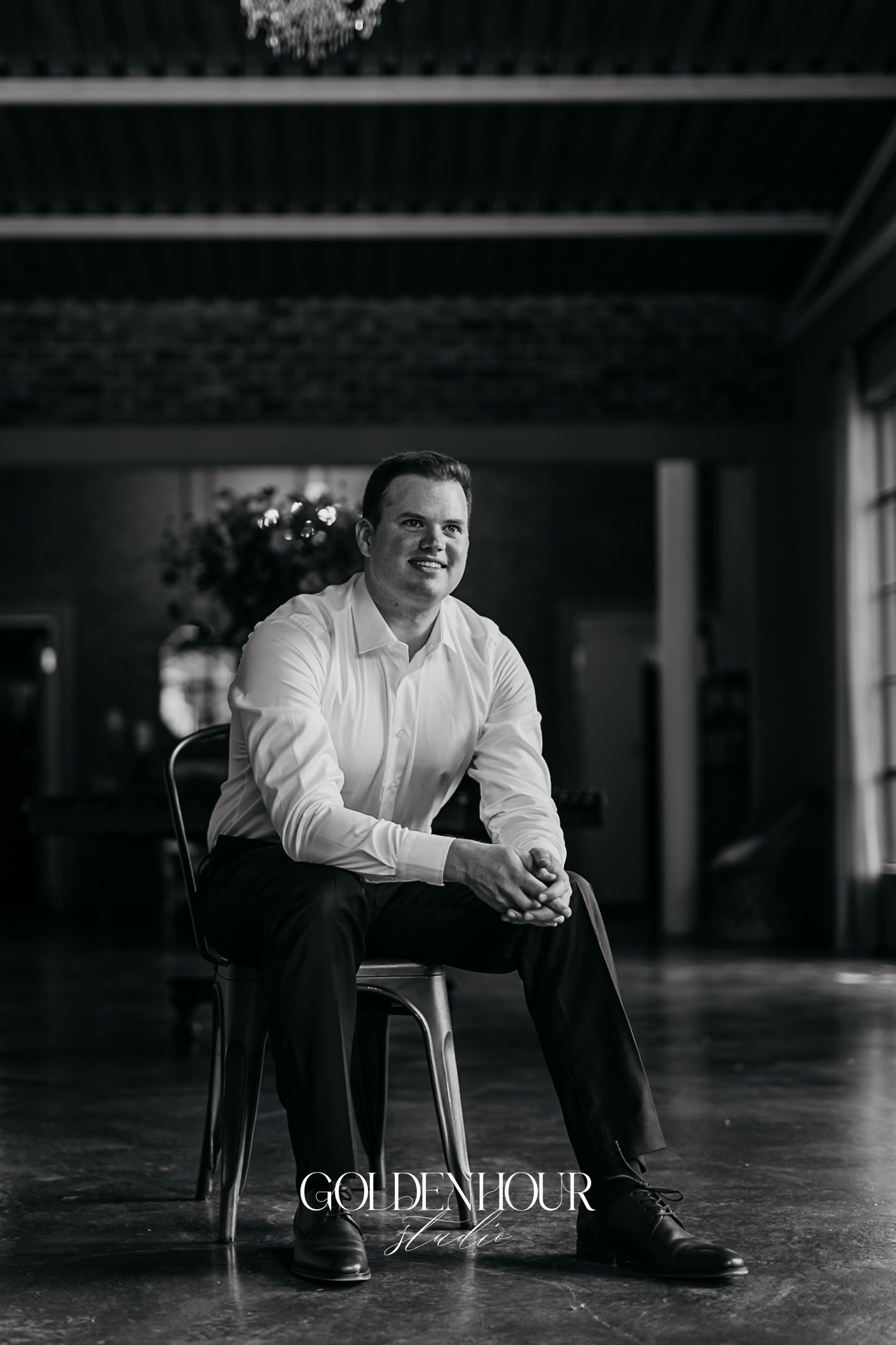 Groom smiling confidently while seated in a chair inside a grand wedding venue, black and white portrait.