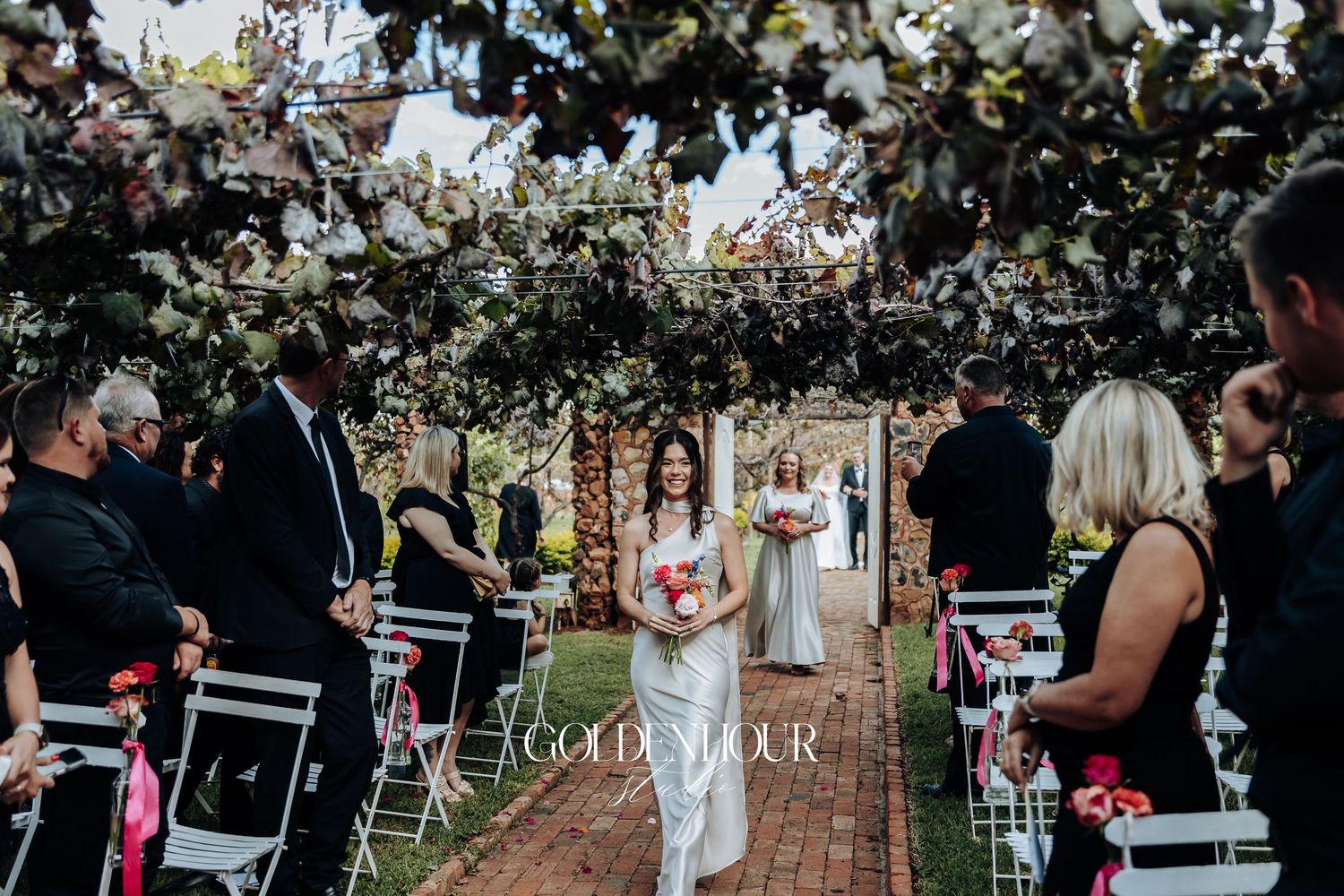Wedding ceremony guests seated in white chairs on a brick patio surrounded by lush greenery and trees.