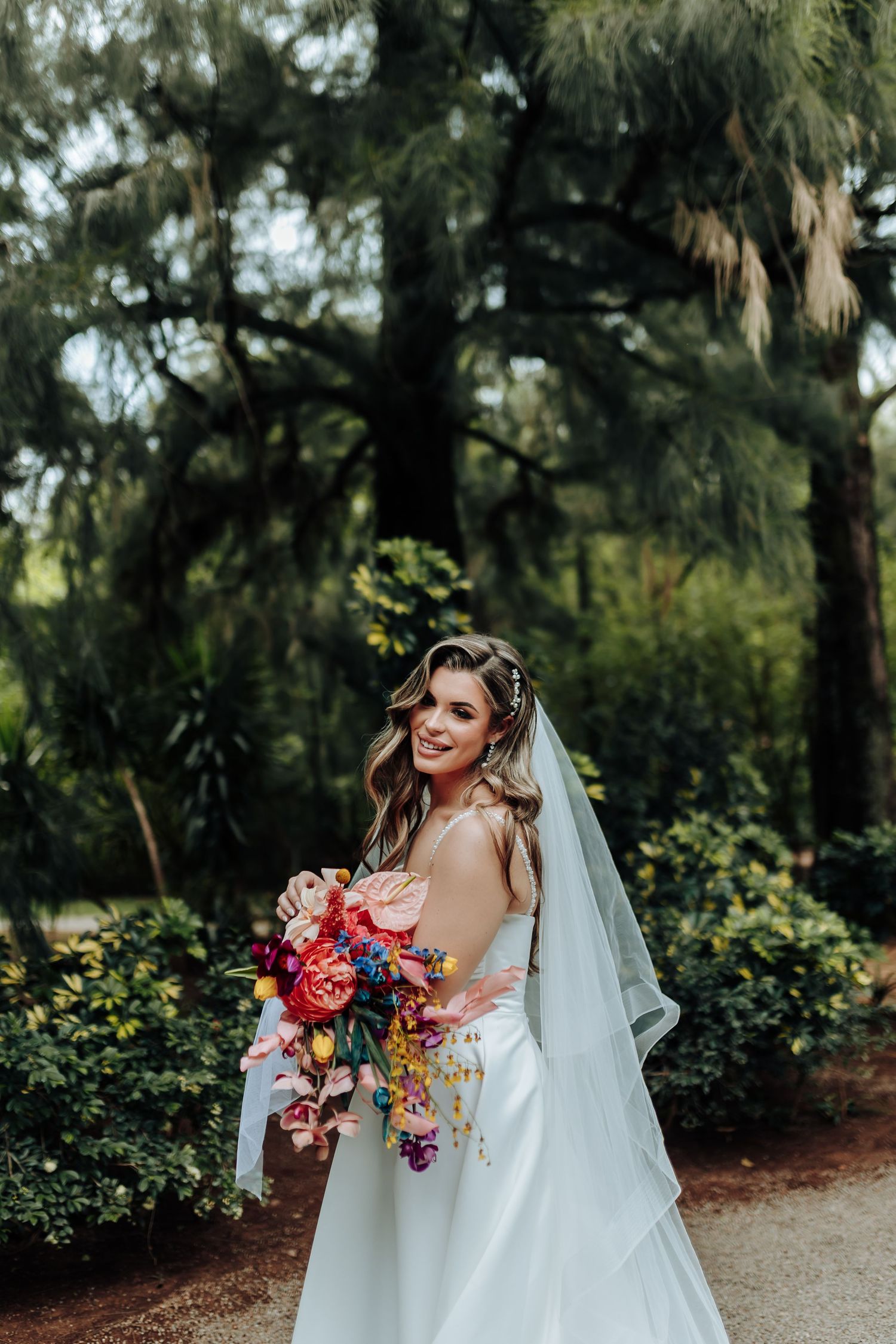 A bride in a white wedding gown holds a colorful bouquet while posing on a garden path.