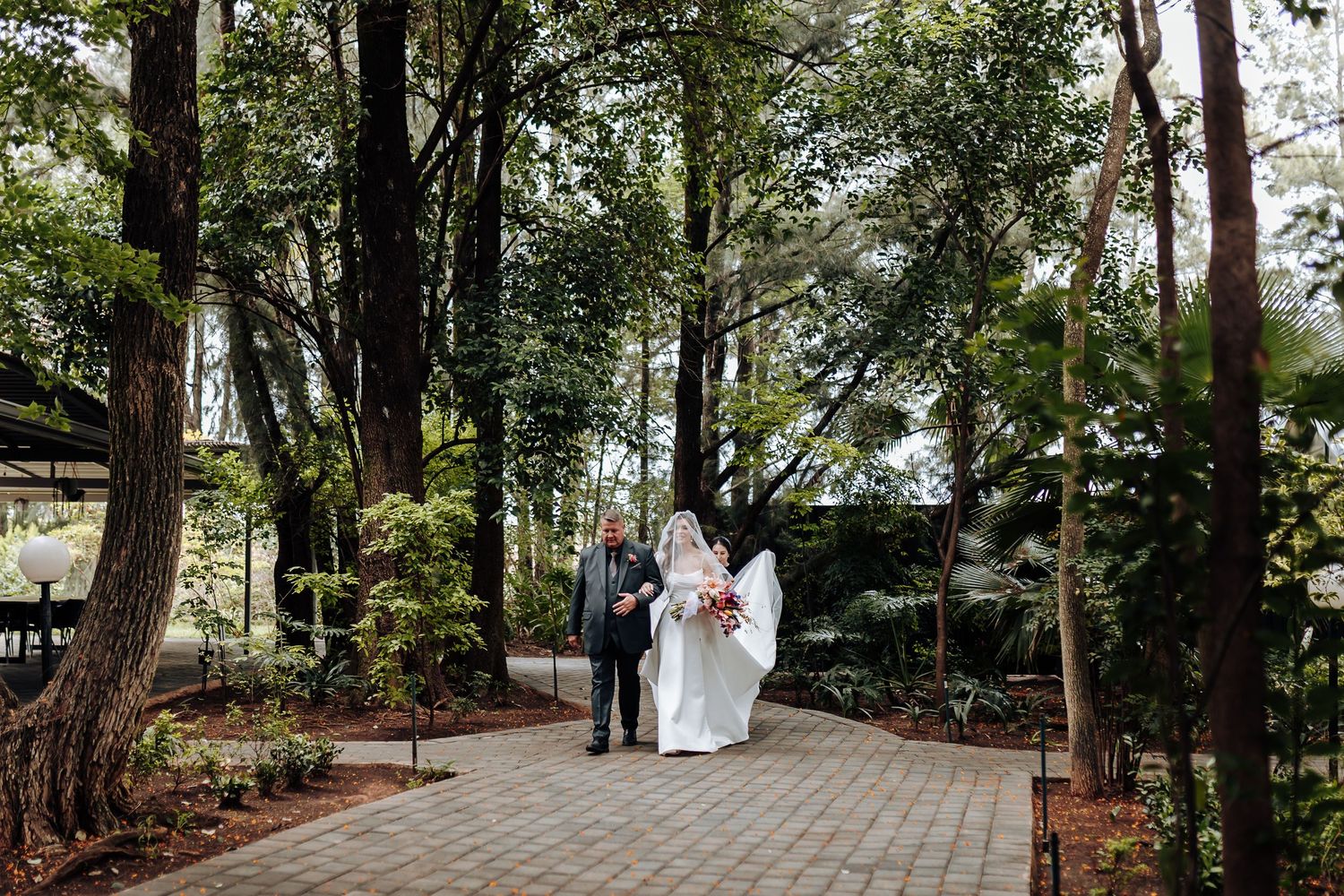 A scenic tree-lined brick pathway leads through a lush garden setting with dappled sunlight filtering through the canopy.