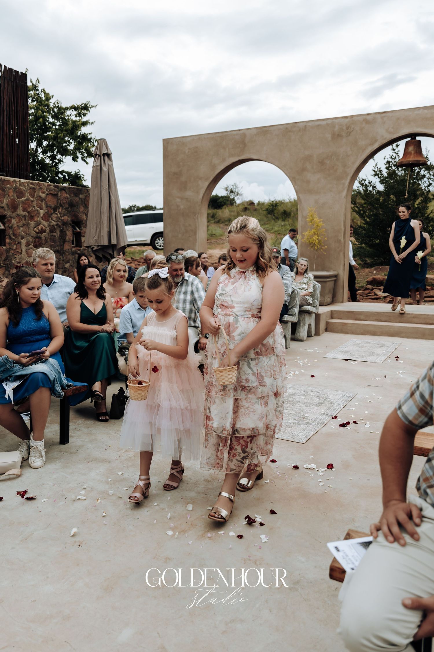Children play and gather rose petals under stone archways at an outdoor wedding venue.