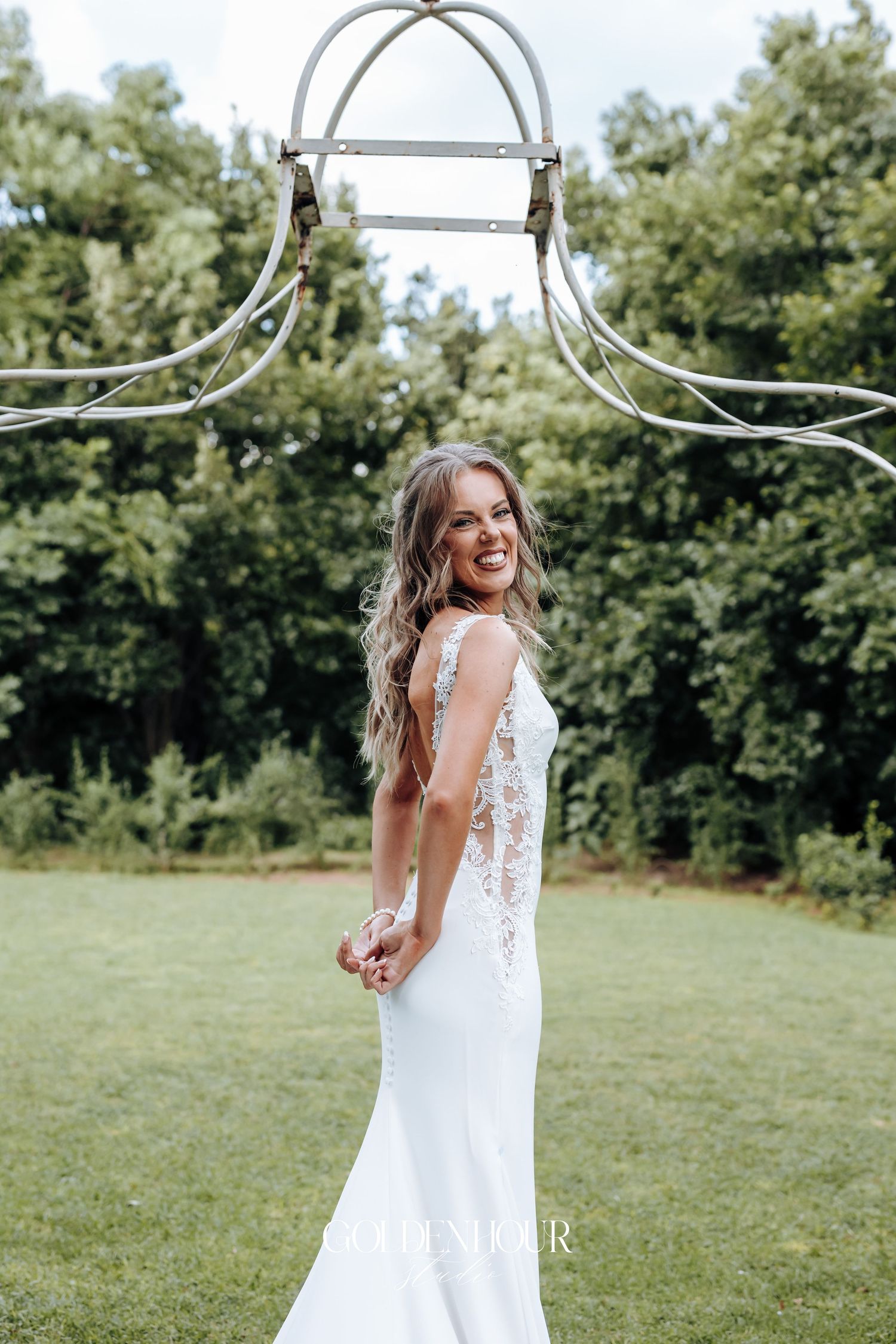 Bride in white lace dress twirls joyfully in an outdoor garden setting with trees in background.
