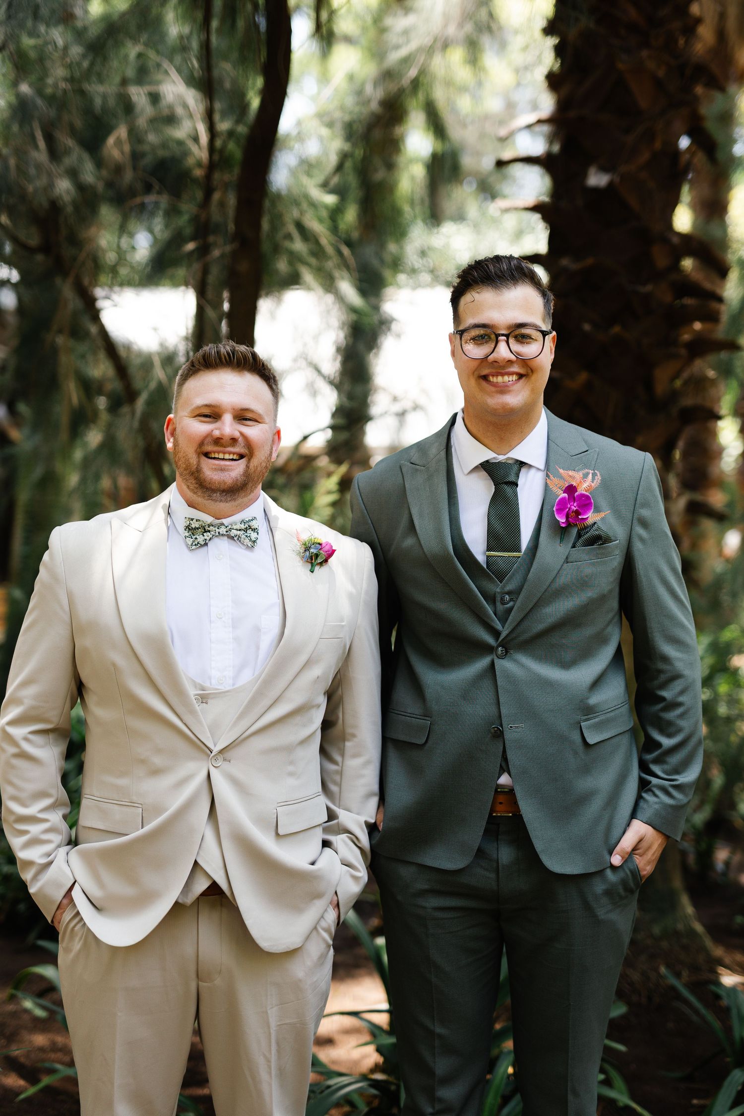 Wedding party members in light beige and dark grey suits pose together in a wooded outdoor setting.