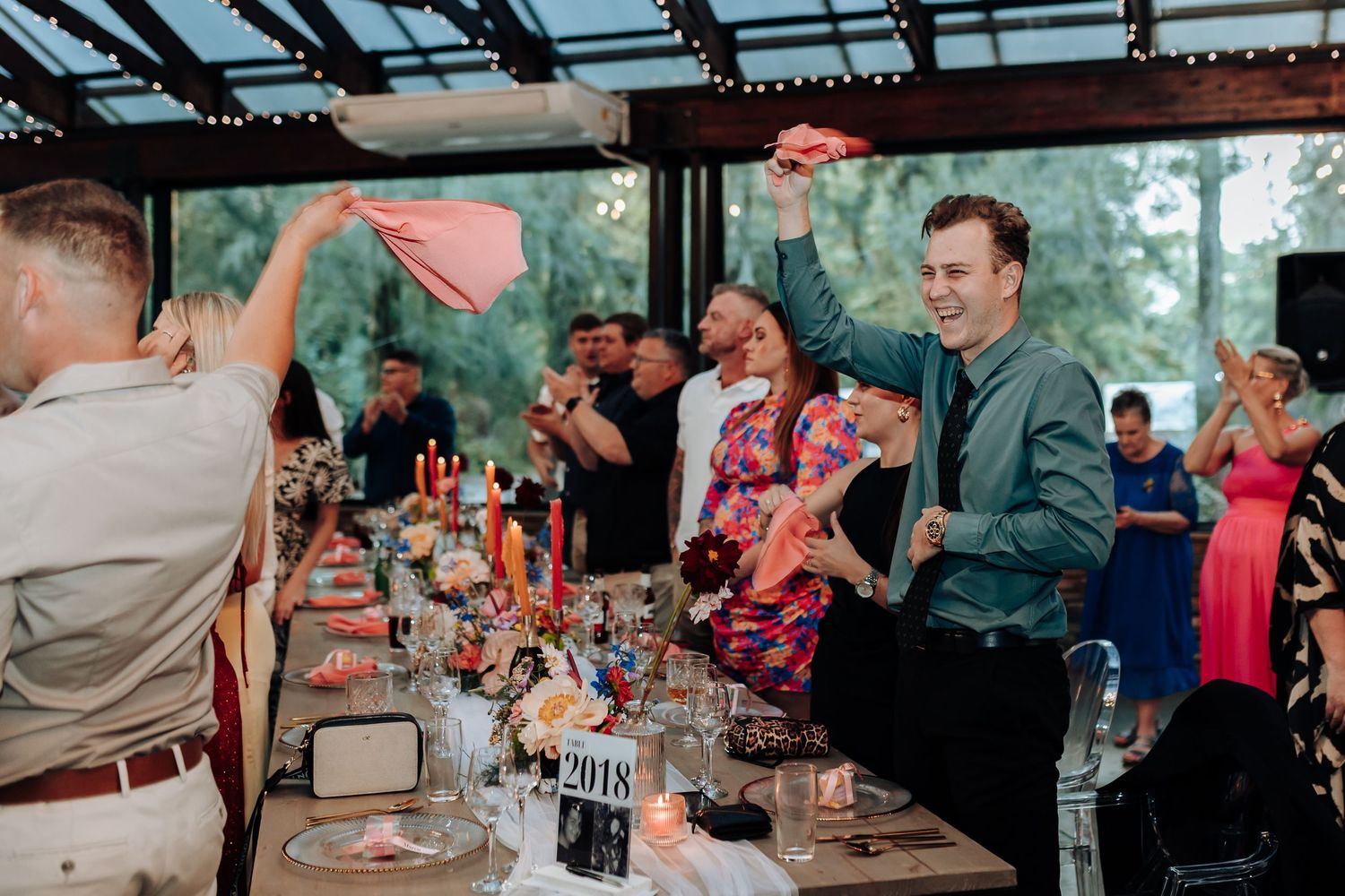 Wedding guests celebrate and dance at a lively reception under a covered outdoor venue with drinks and tables.