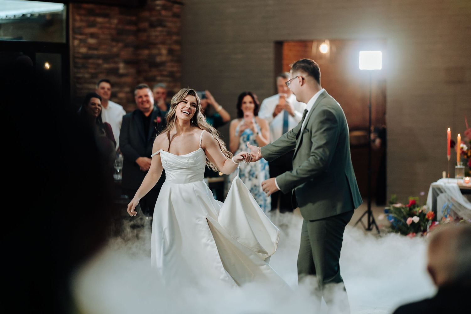 Wedding party dancing in a rustic brick-walled venue with warm lighting and festive atmosphere.