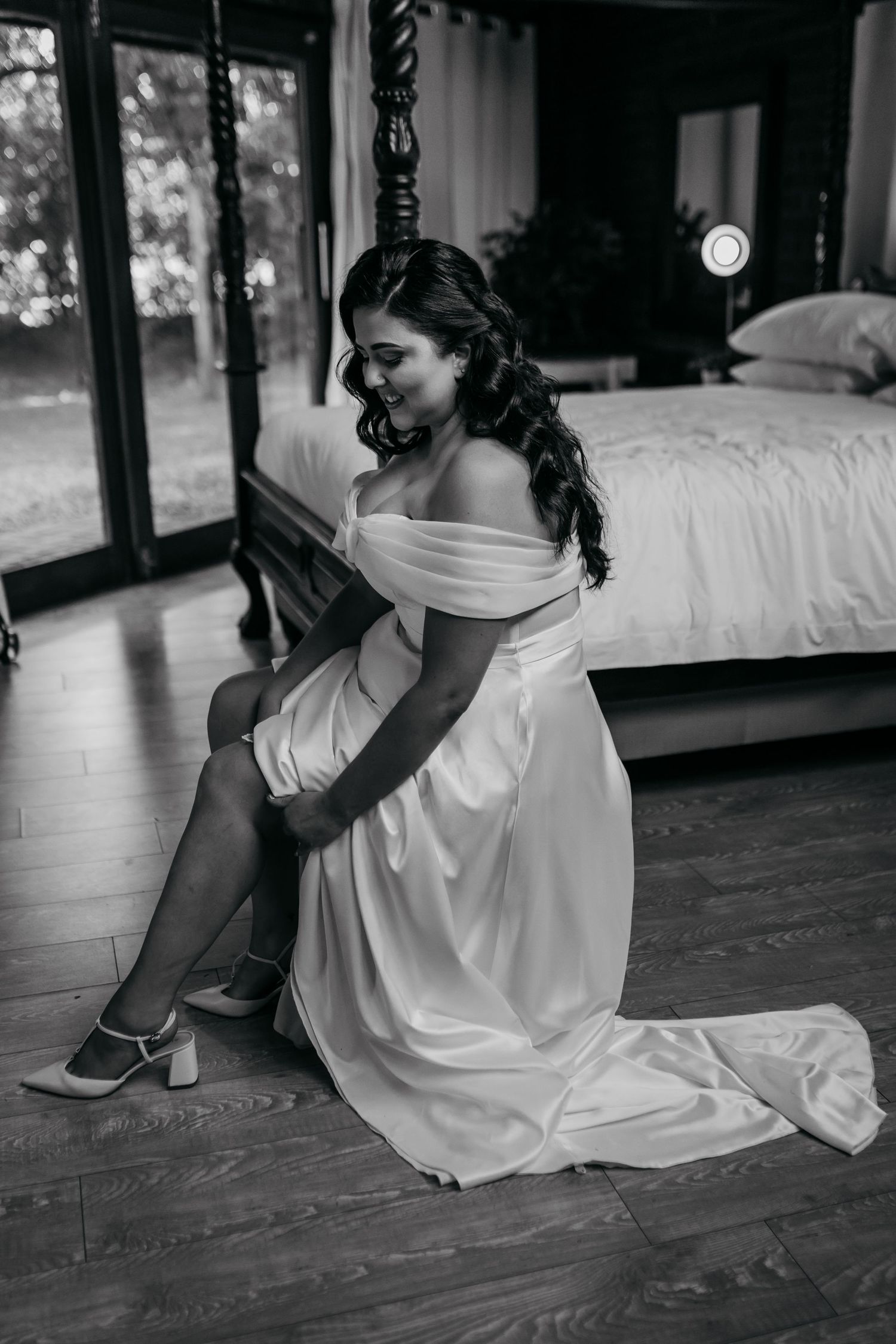 Black and white photo of bride in white dress sitting on wooden floor putting on shoes.