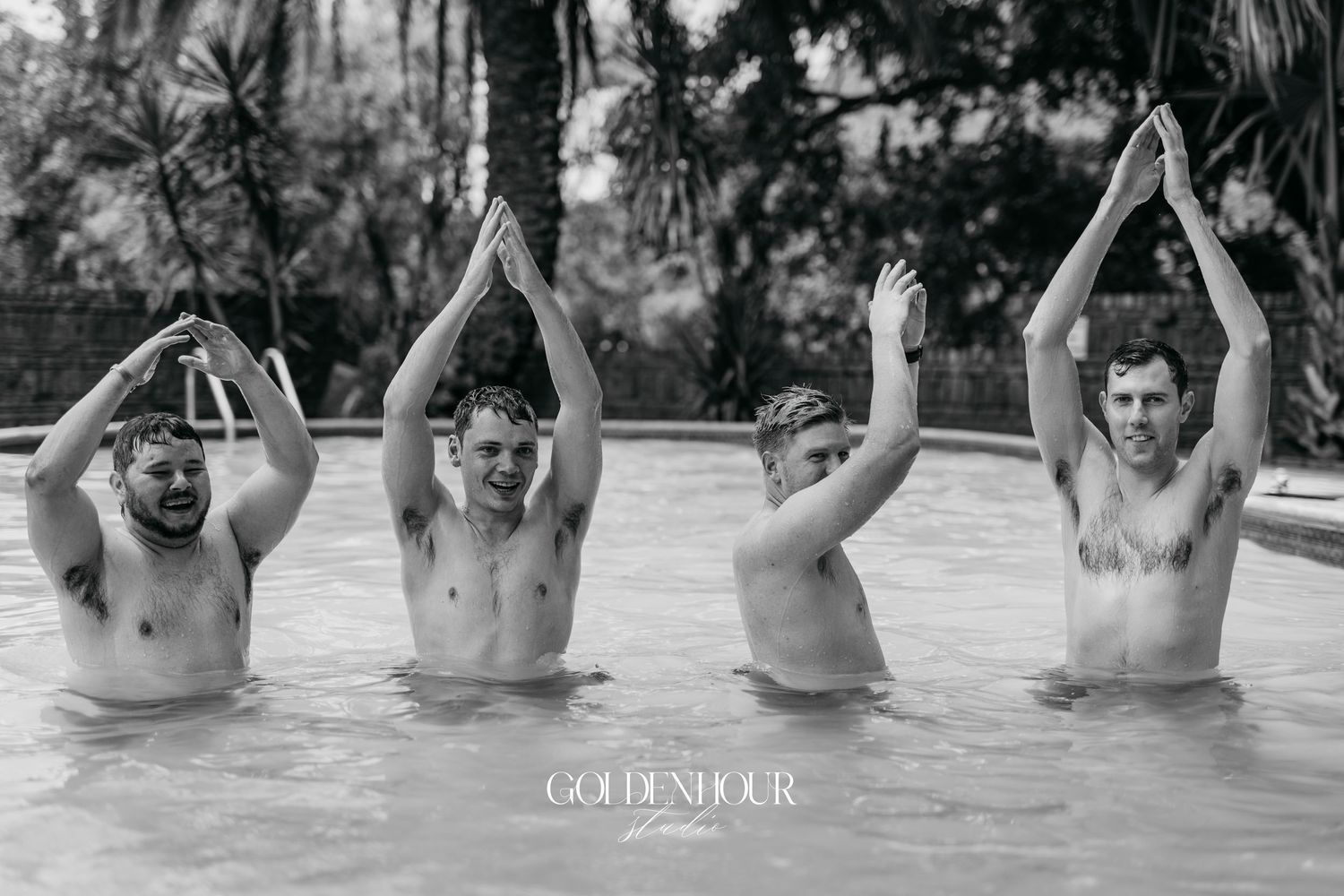 Black and white photo showing multiple people playfully posing in a swimming pool with raised hands.