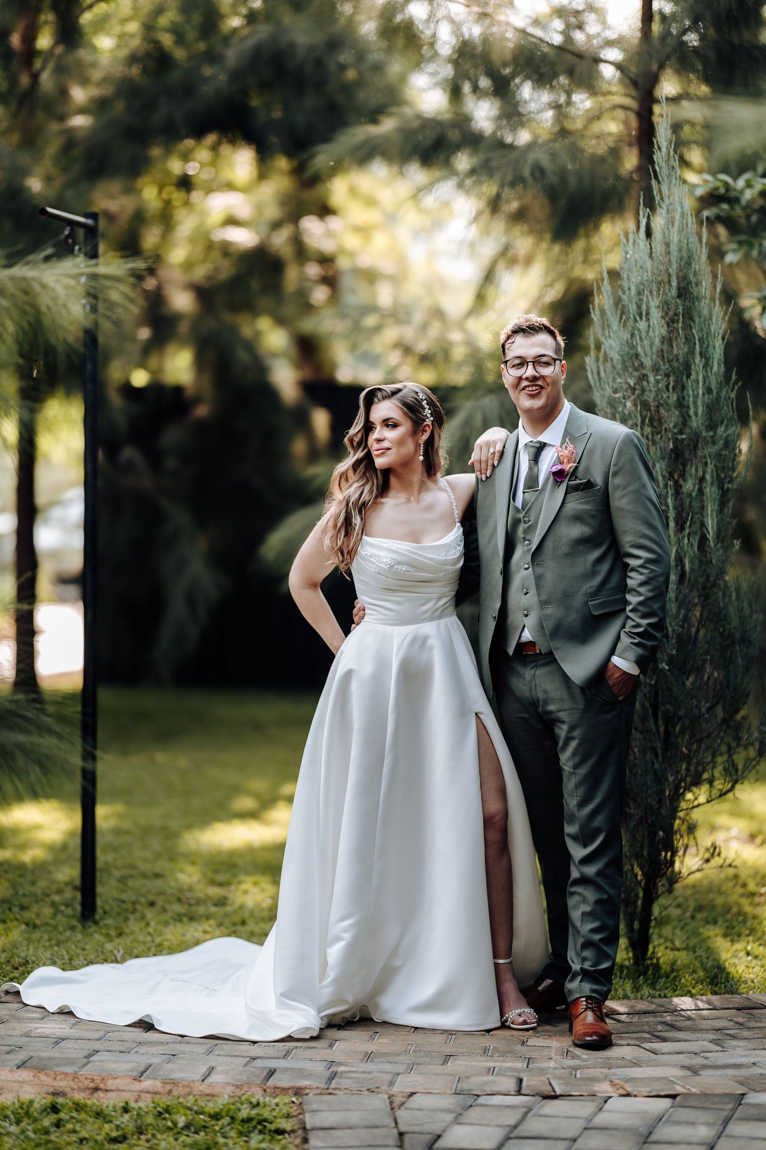 A couple shares a joyful moment together on a stone path surrounded by lush greenery and sunlit trees.