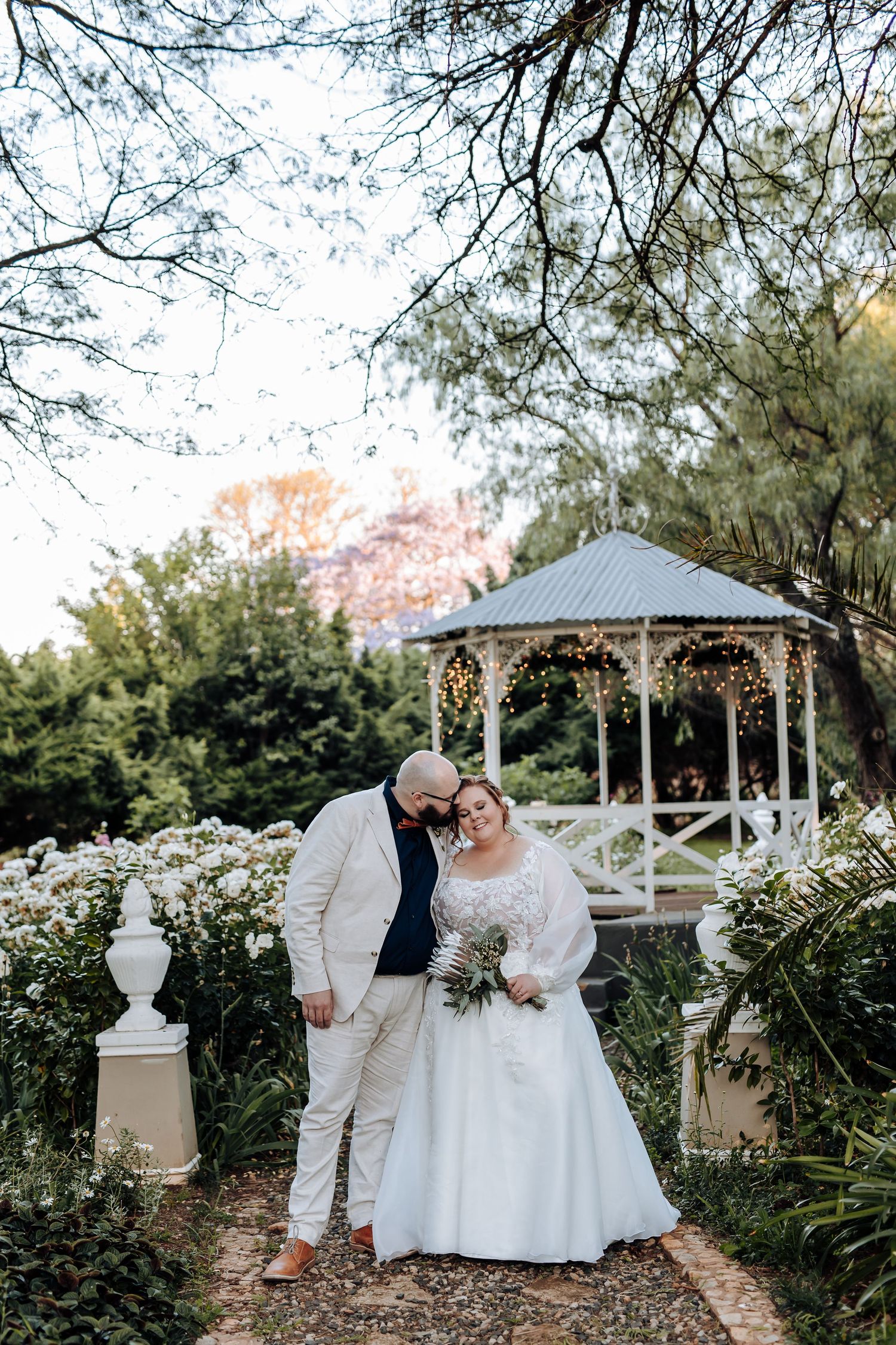A romantic wedding couple poses in front of a white gazebo decorated with string lights in a lush garden setting.