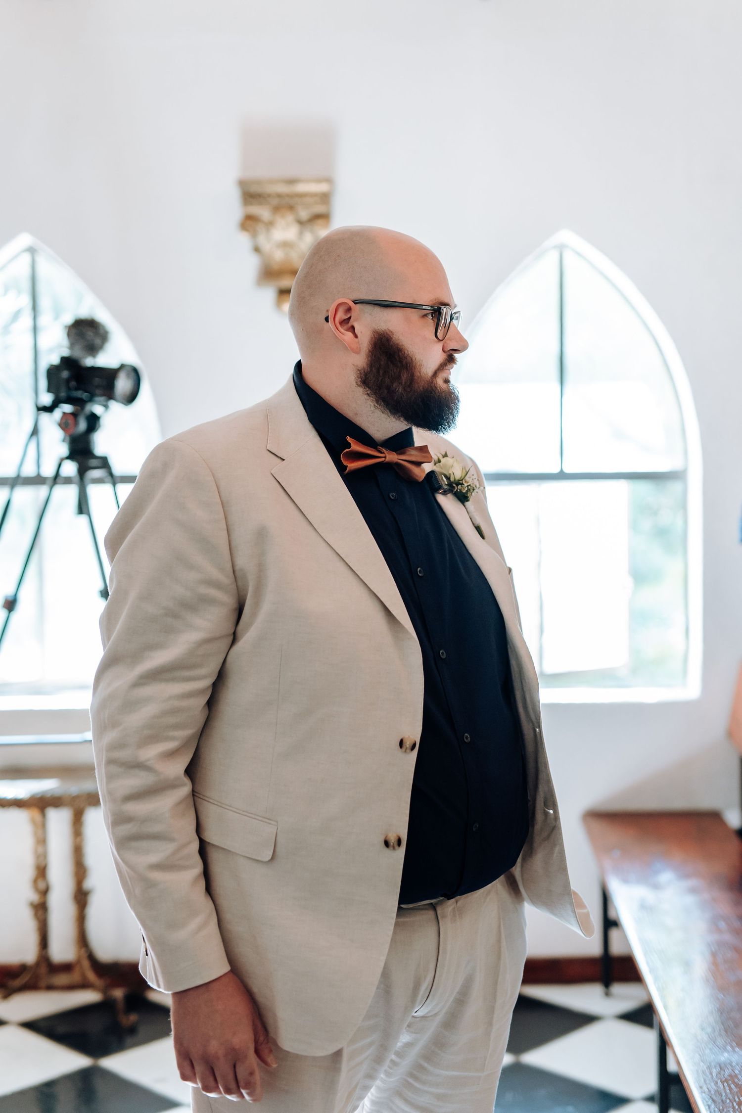 A person in a beige blazer and black shirt stands in a bright white room with a camera visible in the background.