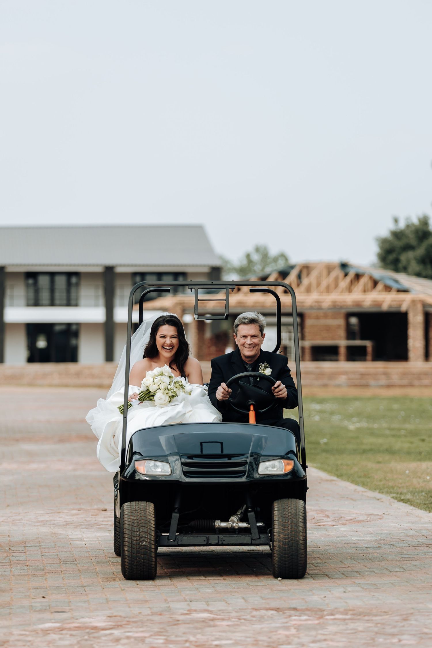A couple rides in a golf cart on their wedding day at a venue with a building and construction site in background.