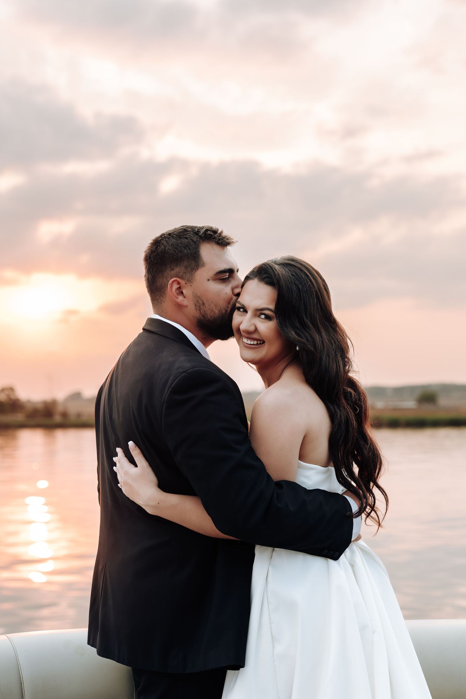 Romantic couple embraces at sunset by a lake, with bride in white dress and groom in black suit.