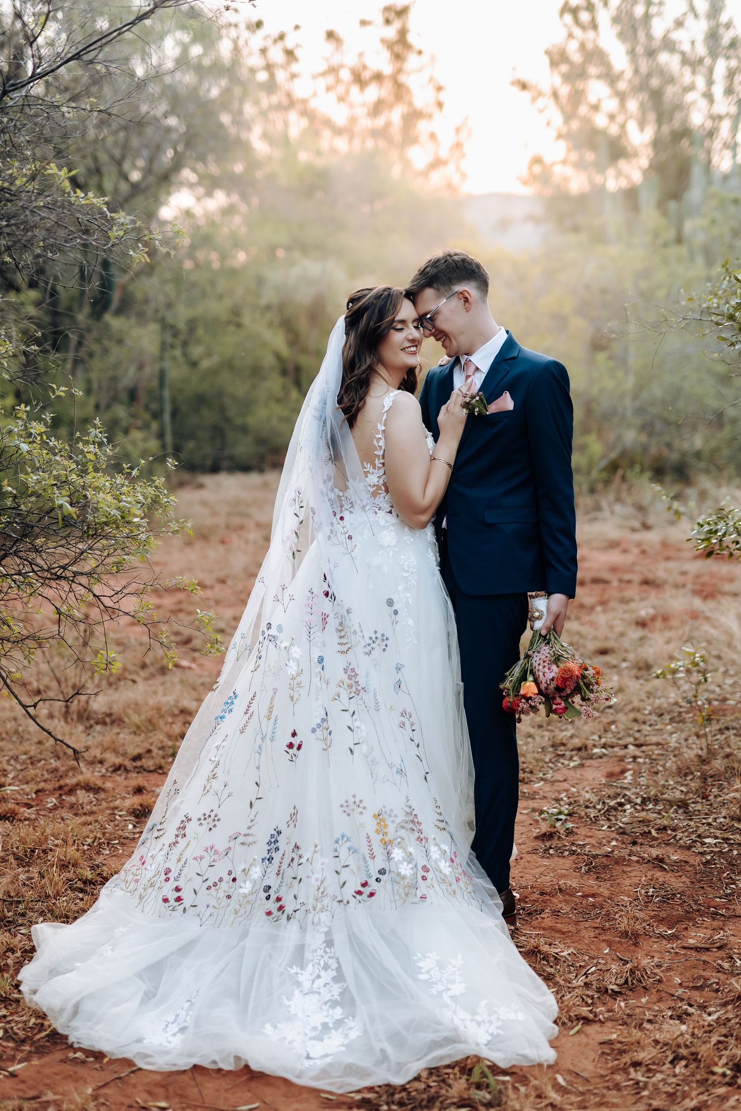 A romantic wedding couple embraces on a rustic dirt path at sunset, with the bride wearing a delicate lace gown.