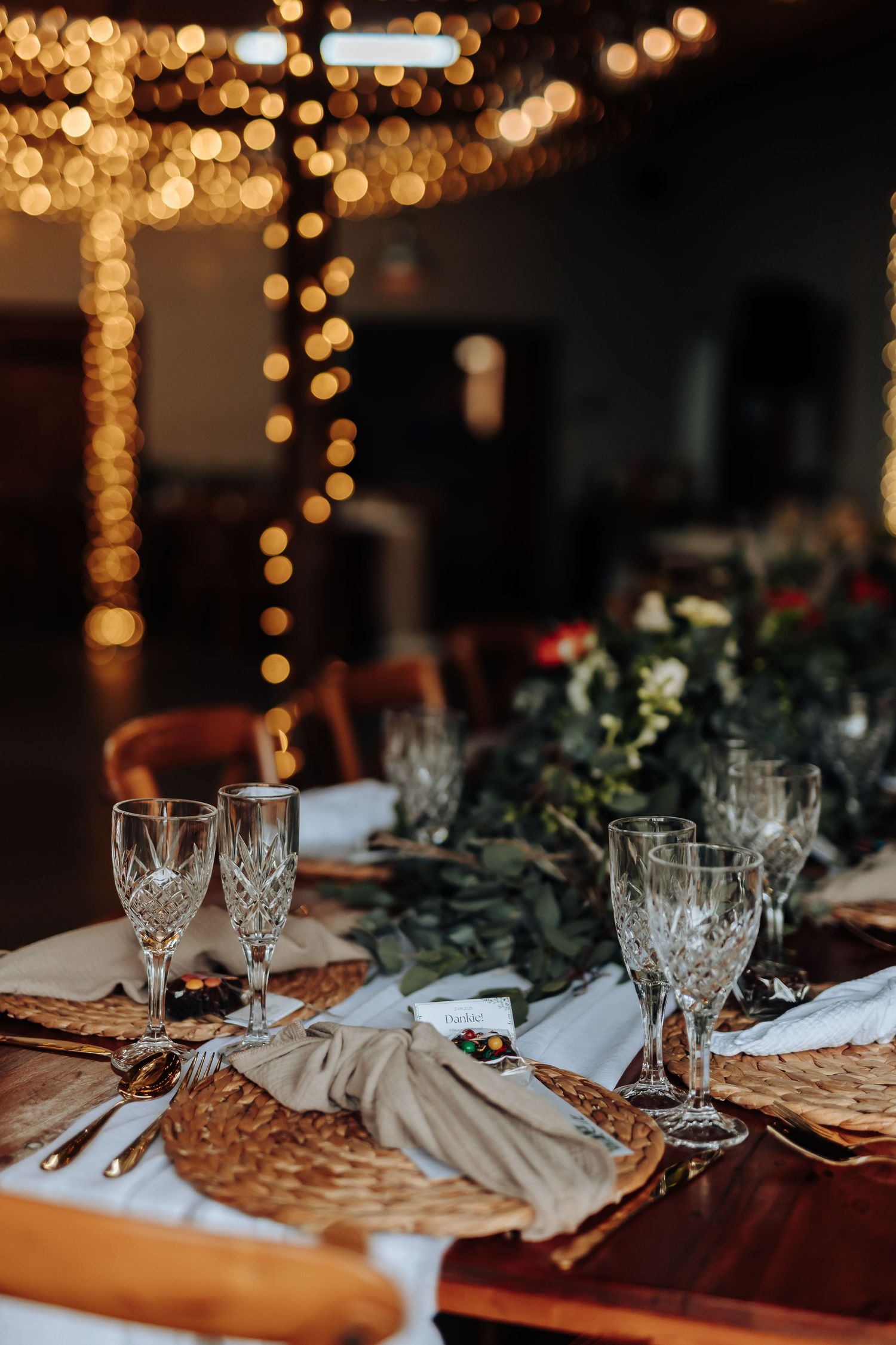 Elegant table setting with crystal glasses and greenery garland under warm string lights creating romantic ambiance.