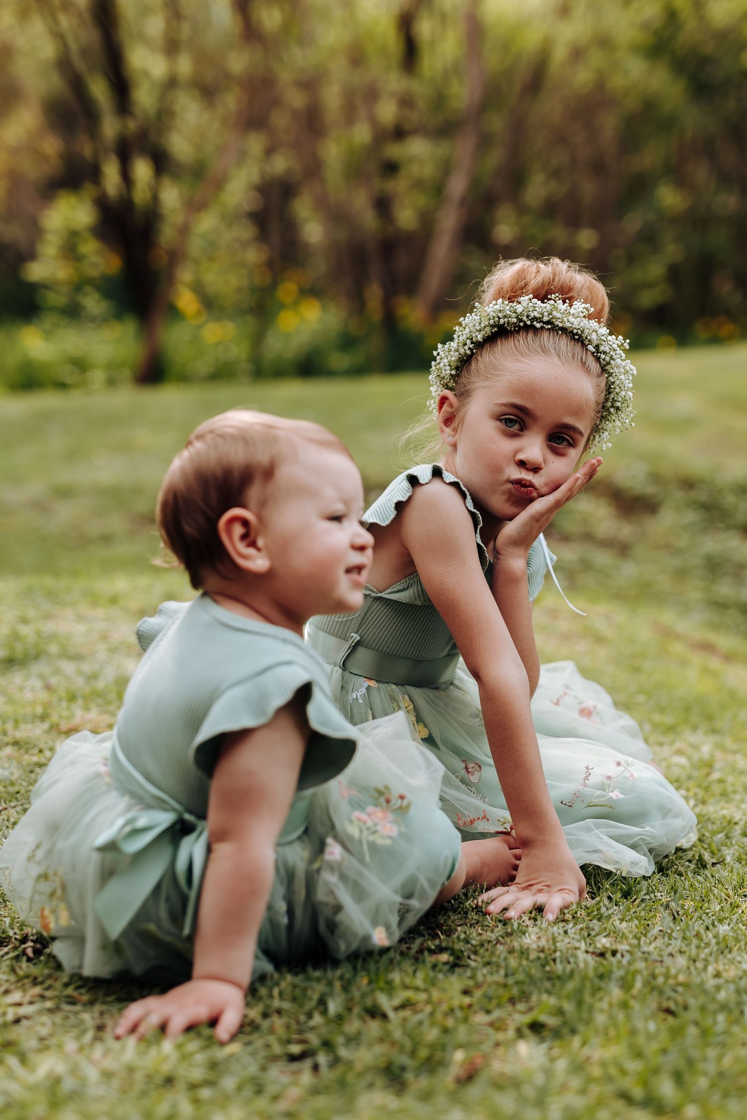 A child in a sage green dress lays on grass wearing a delicate flower crown in their hair.