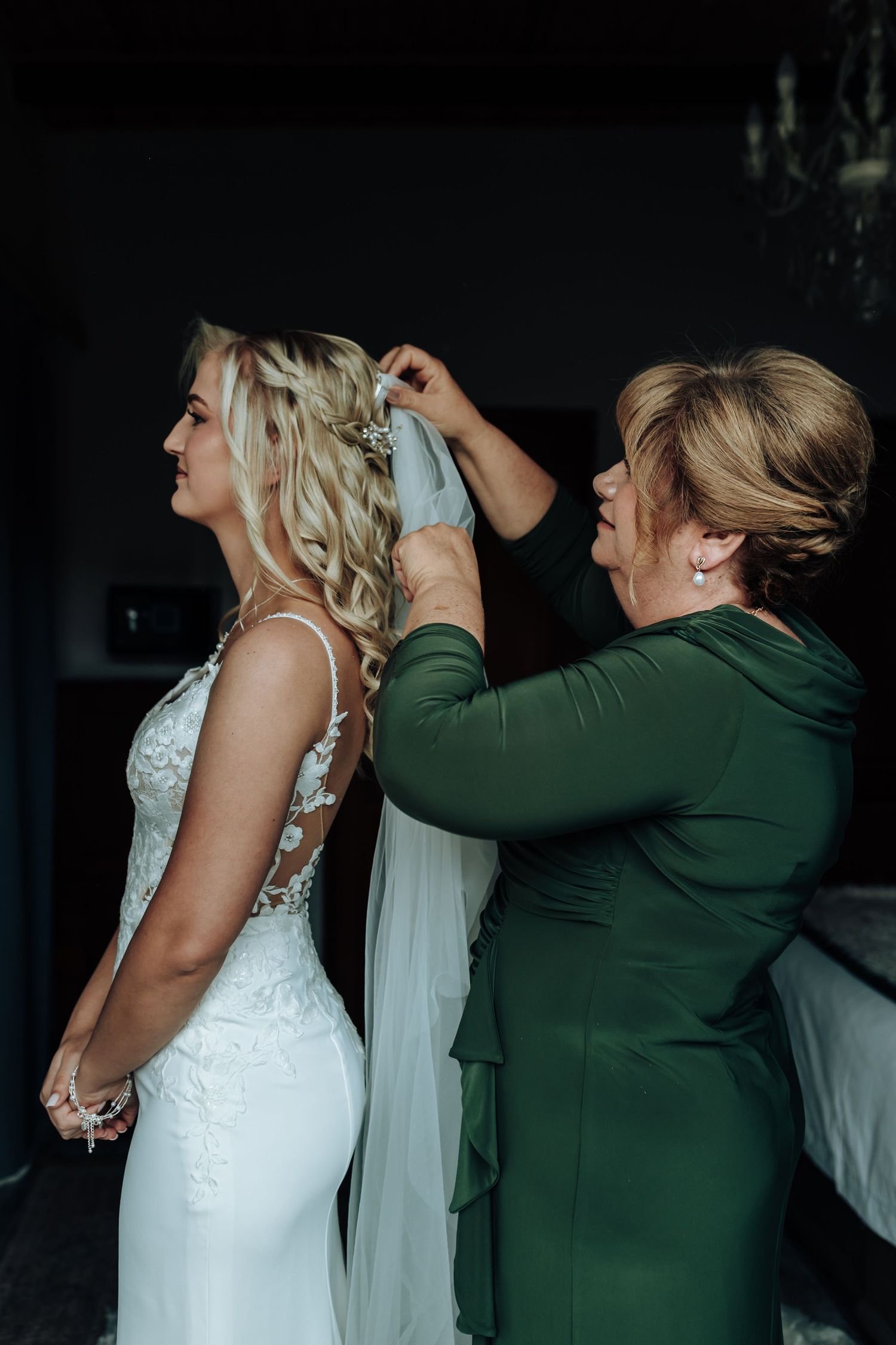 A woman in a white lace wedding dress getting her hair styled by someone wearing an emerald green dress.