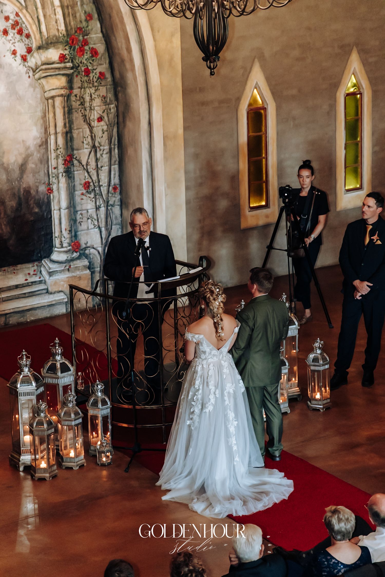Bride and groom stand before officiant at elegant castle chapel wedding ceremony with lanterns and floral mural backdrop.