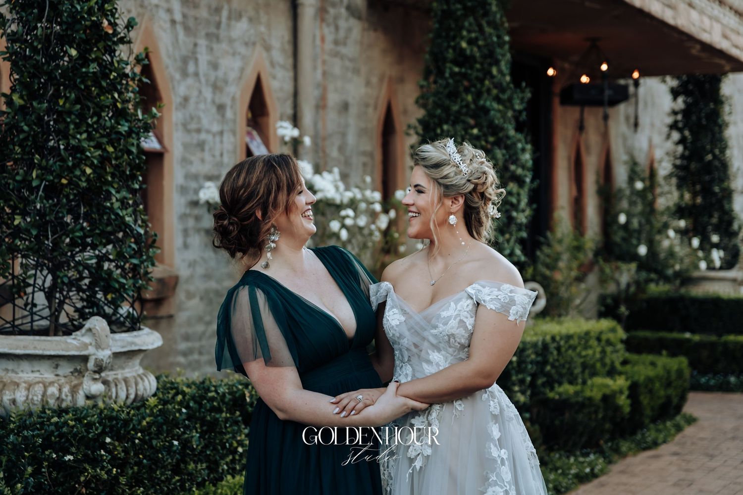 Bride in off-shoulder lace gown smiles with woman in navy dress in a lush garden courtyard setting.