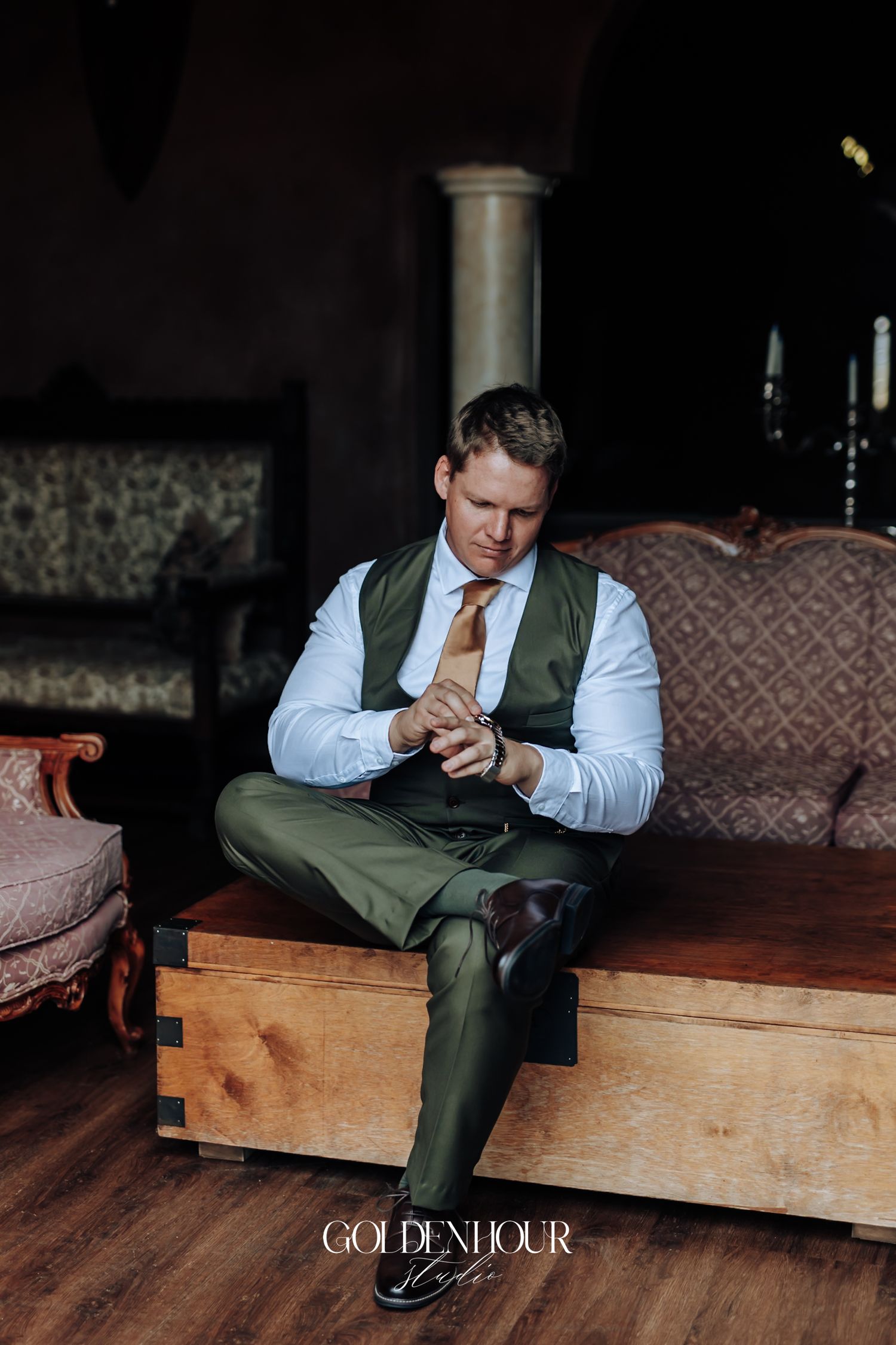 Groom in olive green vest and white shirt adjusting cufflinks while seated on wooden chest in elegant venue.