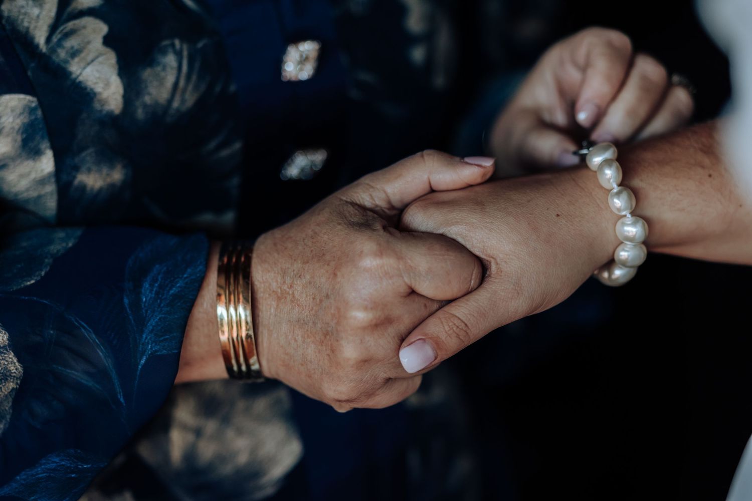 Two elderly hands clasped together, one wearing gold bangles and the other a pearl bracelet, sharing a tender moment.