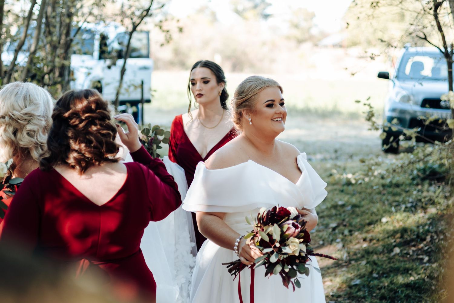 Bride in white off-shoulder gown holds bouquet with bridesmaids in red dresses at outdoor wedding ceremony.