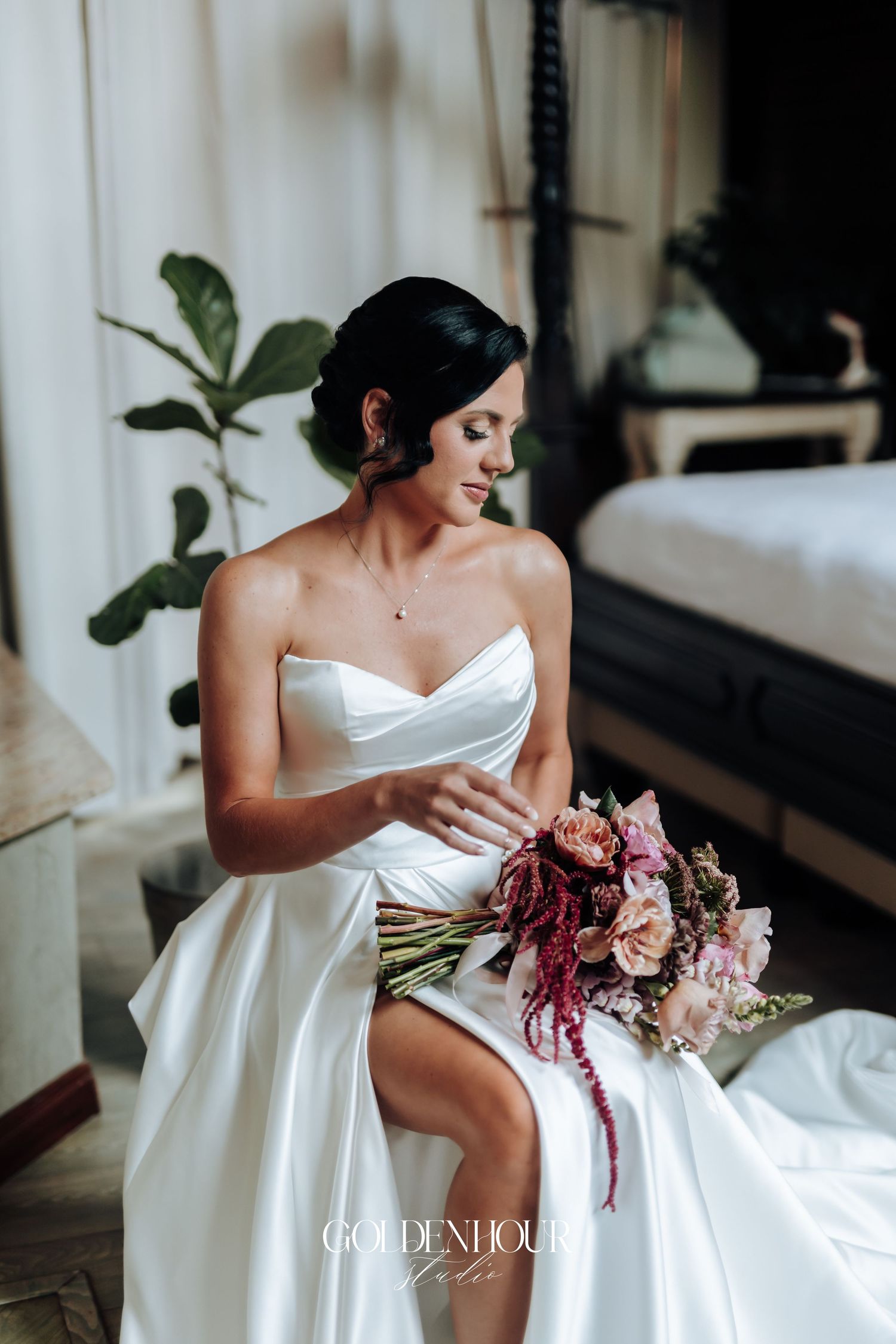 Bride in strapless white gown seated holding pink floral bouquet with amaranthus in elegant indoor setting.