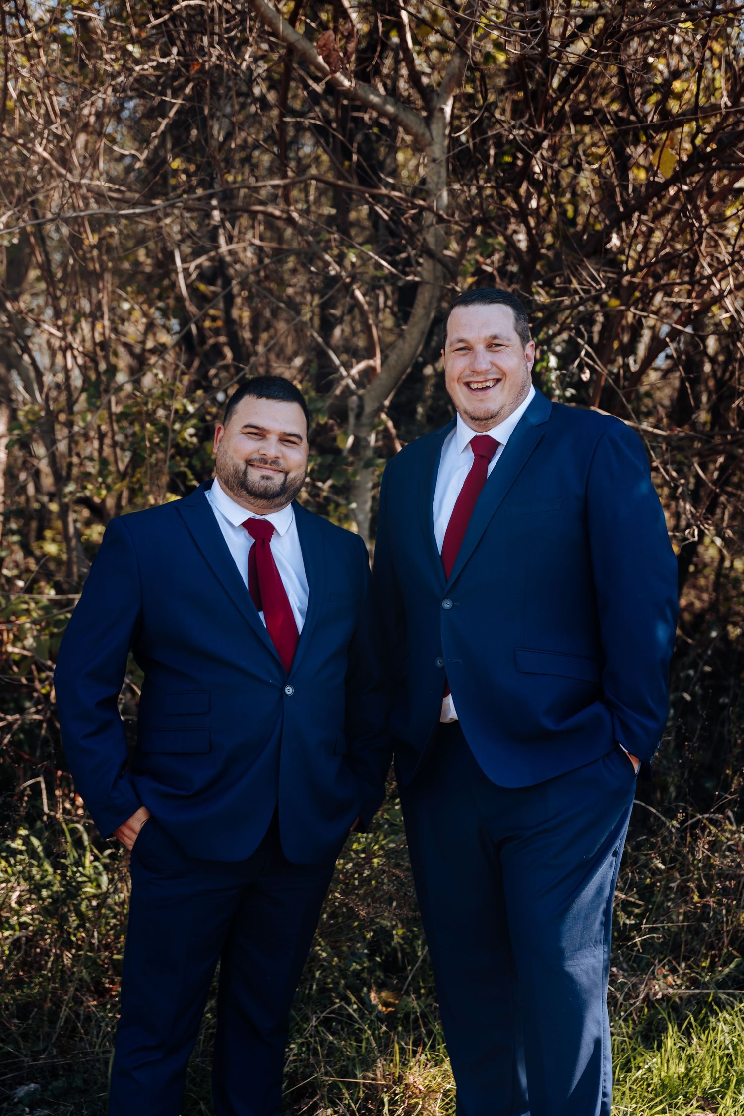 Two men in matching navy suits and red ties smiling outdoors in a natural setting.