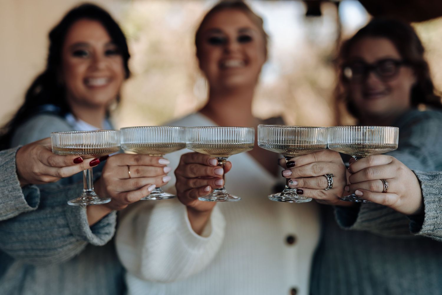 Three women smiling and toasting with elegant coupe glasses filled with cocktails at a celebration.