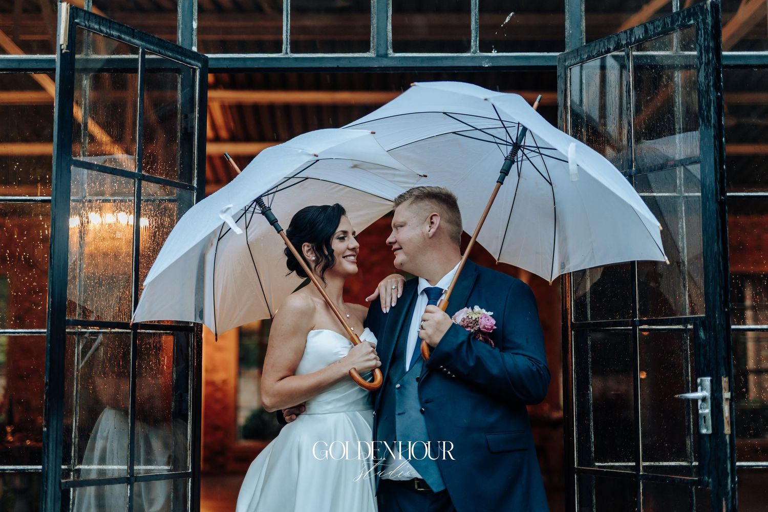 Bride and groom sharing romantic moment under white umbrella outside glass doors on rainy wedding day.