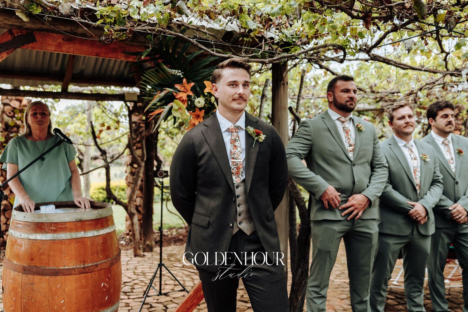 Wedding party in sage green and dark suits standing together by rustic wine barrels.