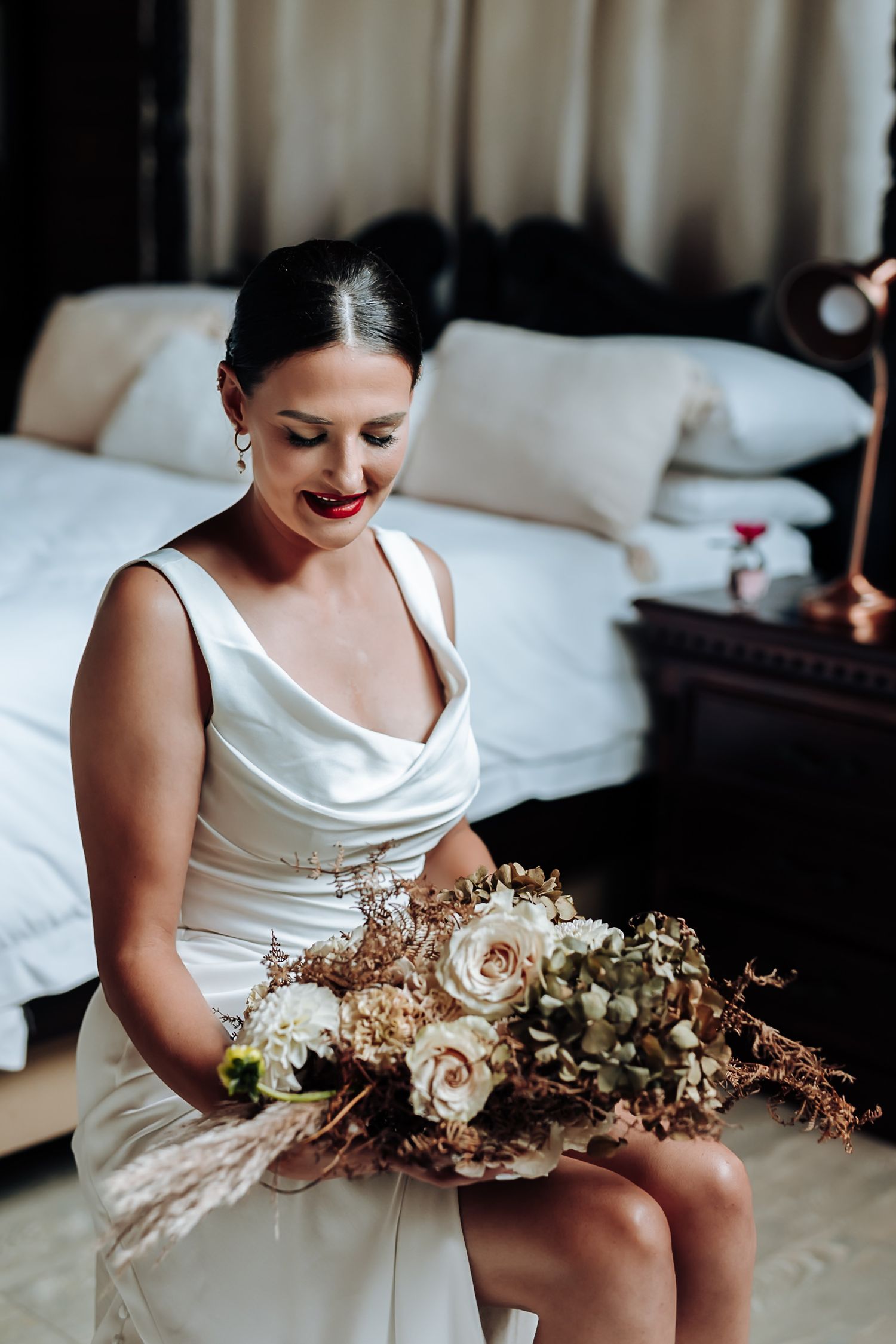 A bride in white satin holds a romantic dried floral arrangement in neutral tones.