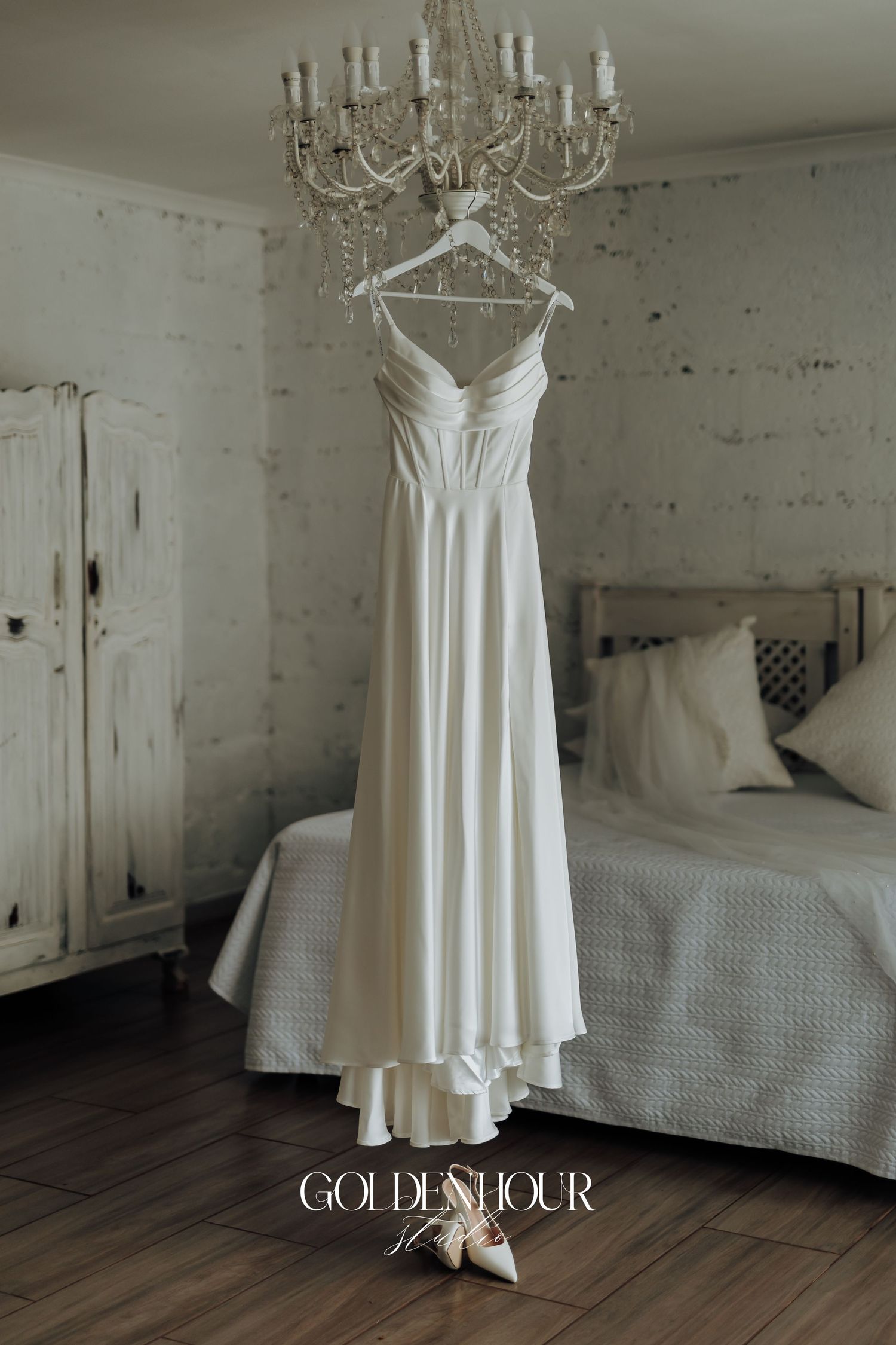 Elegant white wedding dress hanging in a rustic bedroom with chandelier and wooden floors.