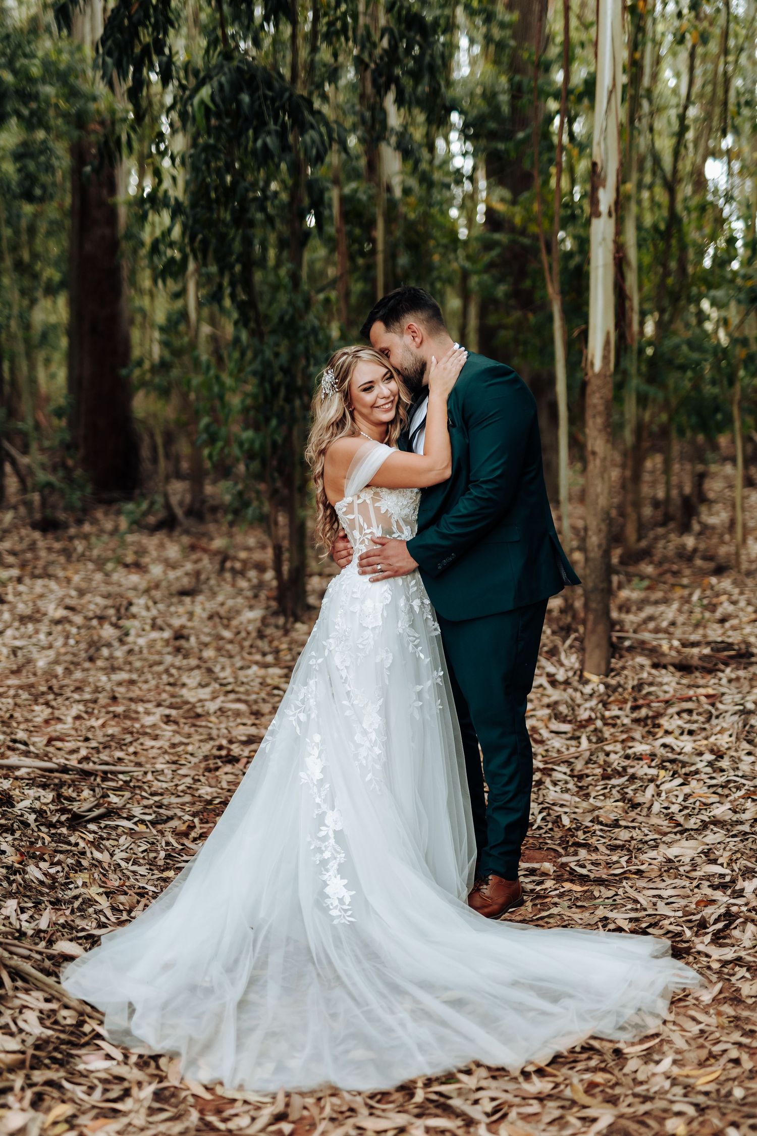 A romantic wedding couple embraces in a serene eucalyptus forest with a flowing white lace gown trailing behind them.