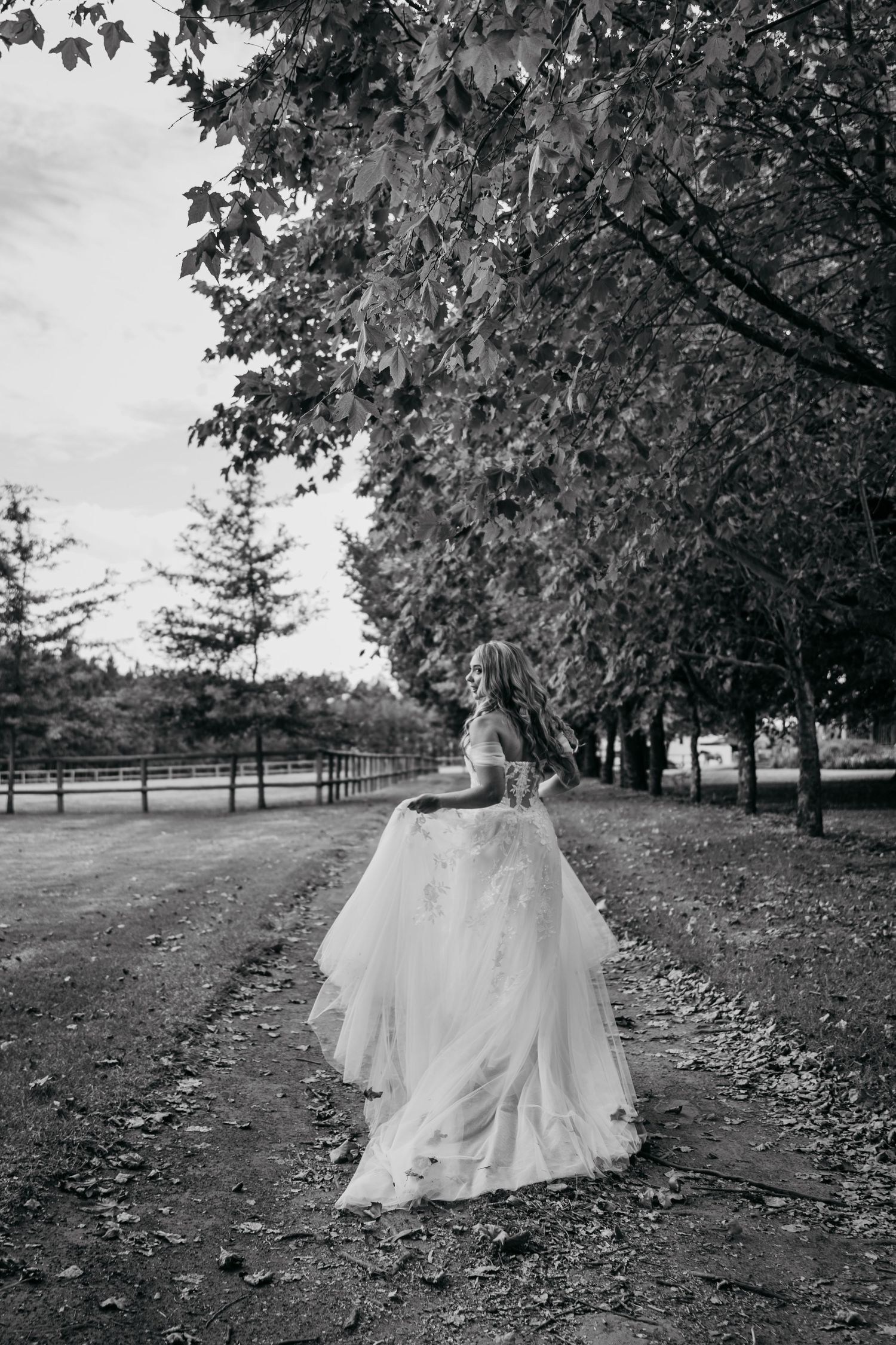 A bride in a flowing white gown walks along a tree-lined path at a rustic farm venue in black and white photography.