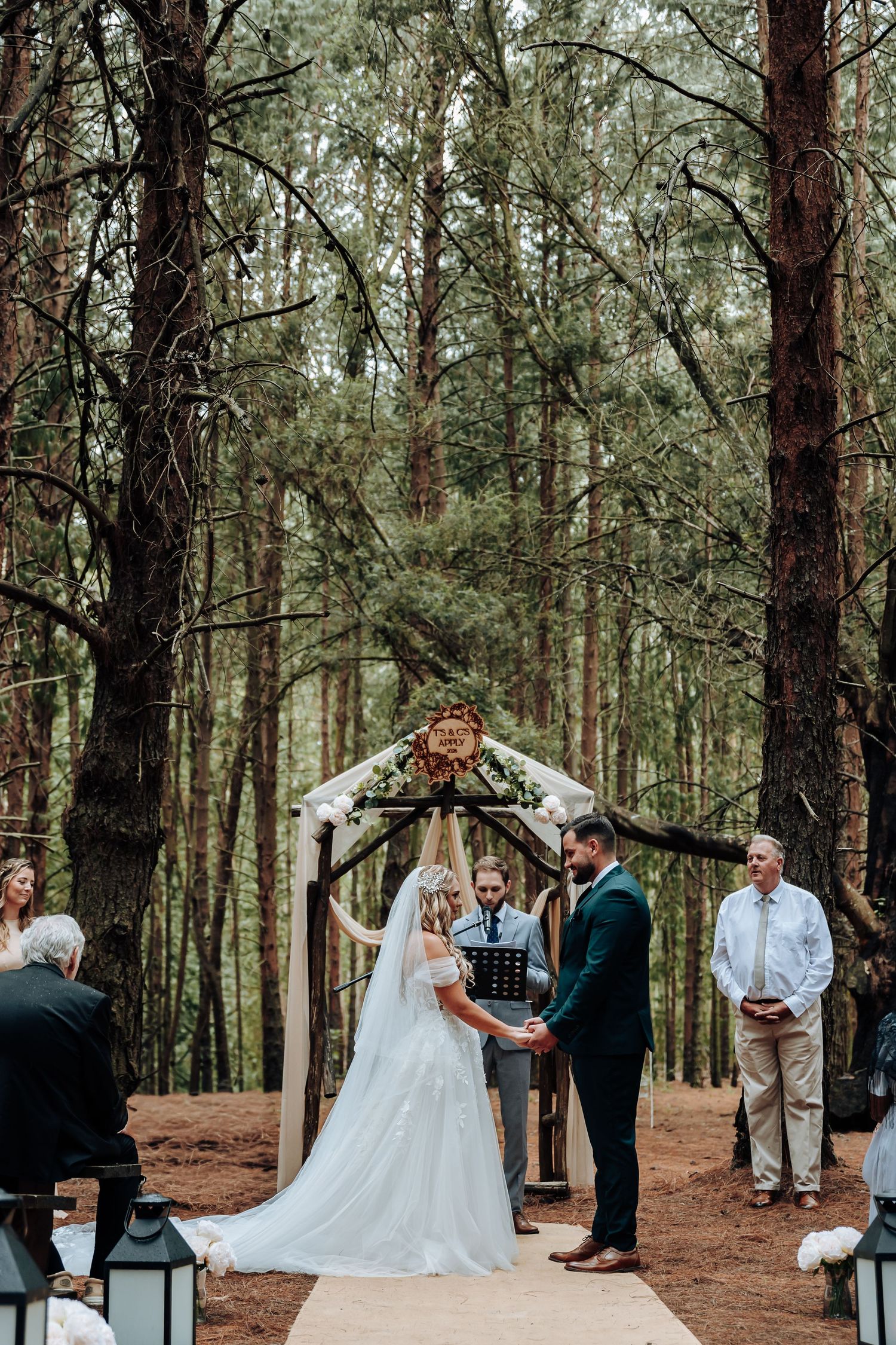 A wedding ceremony takes place in a serene pine forest setting with guests gathered around the couple.