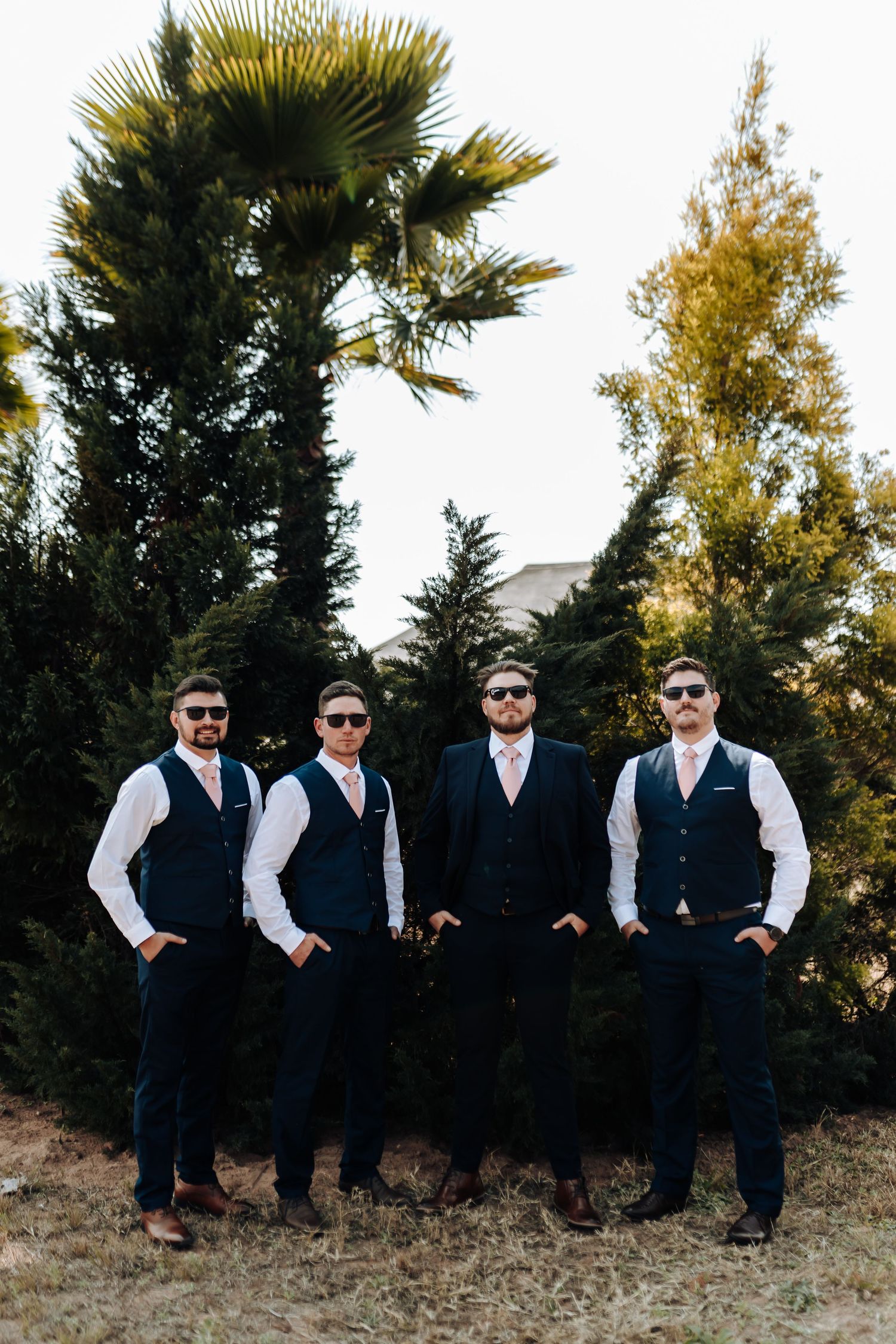 Groomsmen in navy vests and sunglasses pose casually against a backdrop of palm trees and autumn foliage.