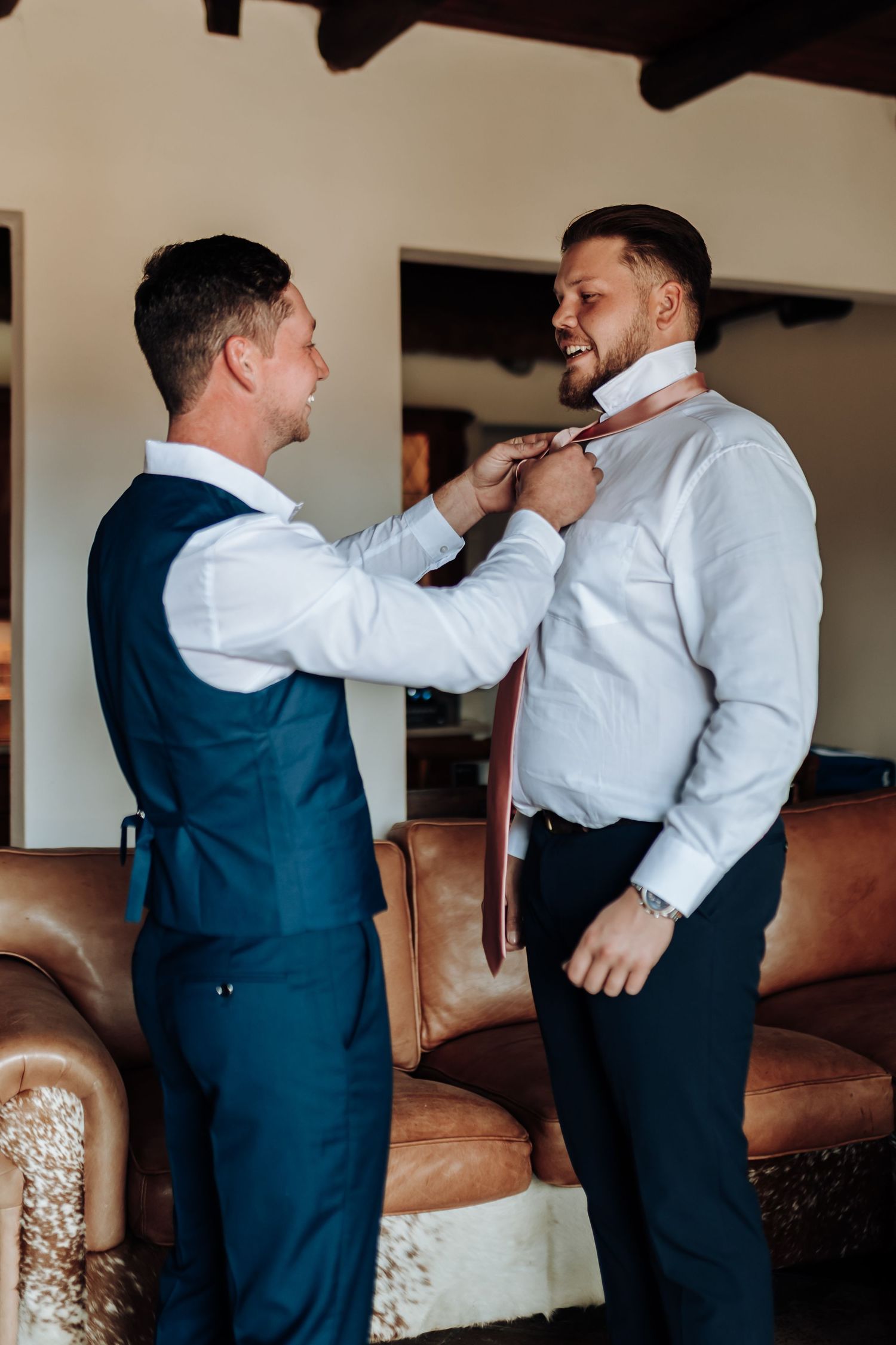 Two men in formal attire help each other with neckties while sharing a lighthearted moment near a brown leather couch.