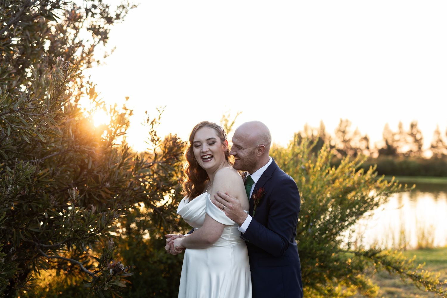 Bride and groom embrace at golden hour sunset near a pond, surrounded by lush greenery on their wedding day.