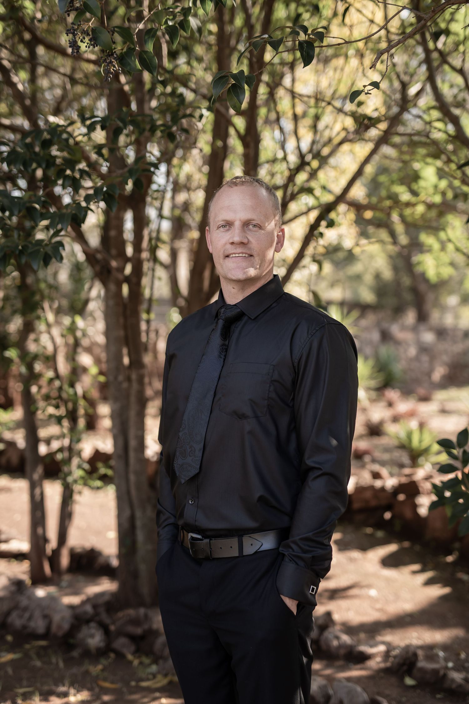 Professional portrait of a person in formal black attire standing outdoors against a natural wooded background.