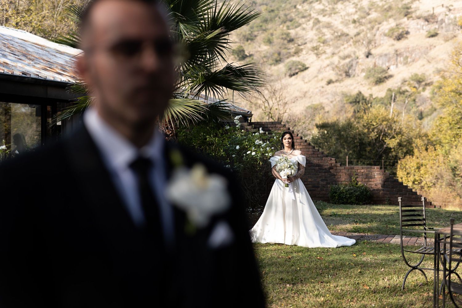 A wedding scene in a garden shows a couple in formal attire during a traditional first look moment.