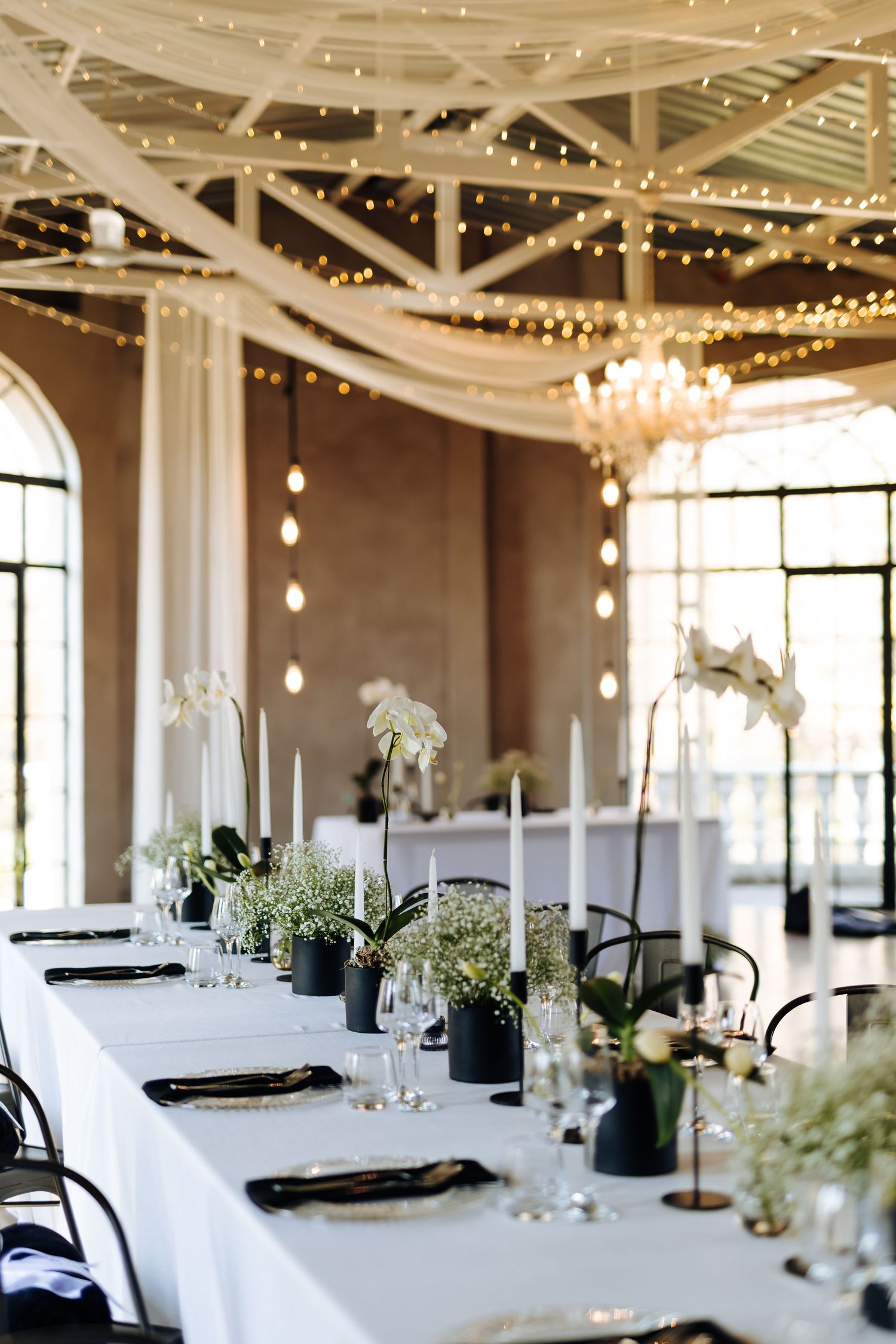 Elegant wedding reception table with white linens, ostrich feather centerpieces, and twinkle lights draped from exposed ceiling beams.