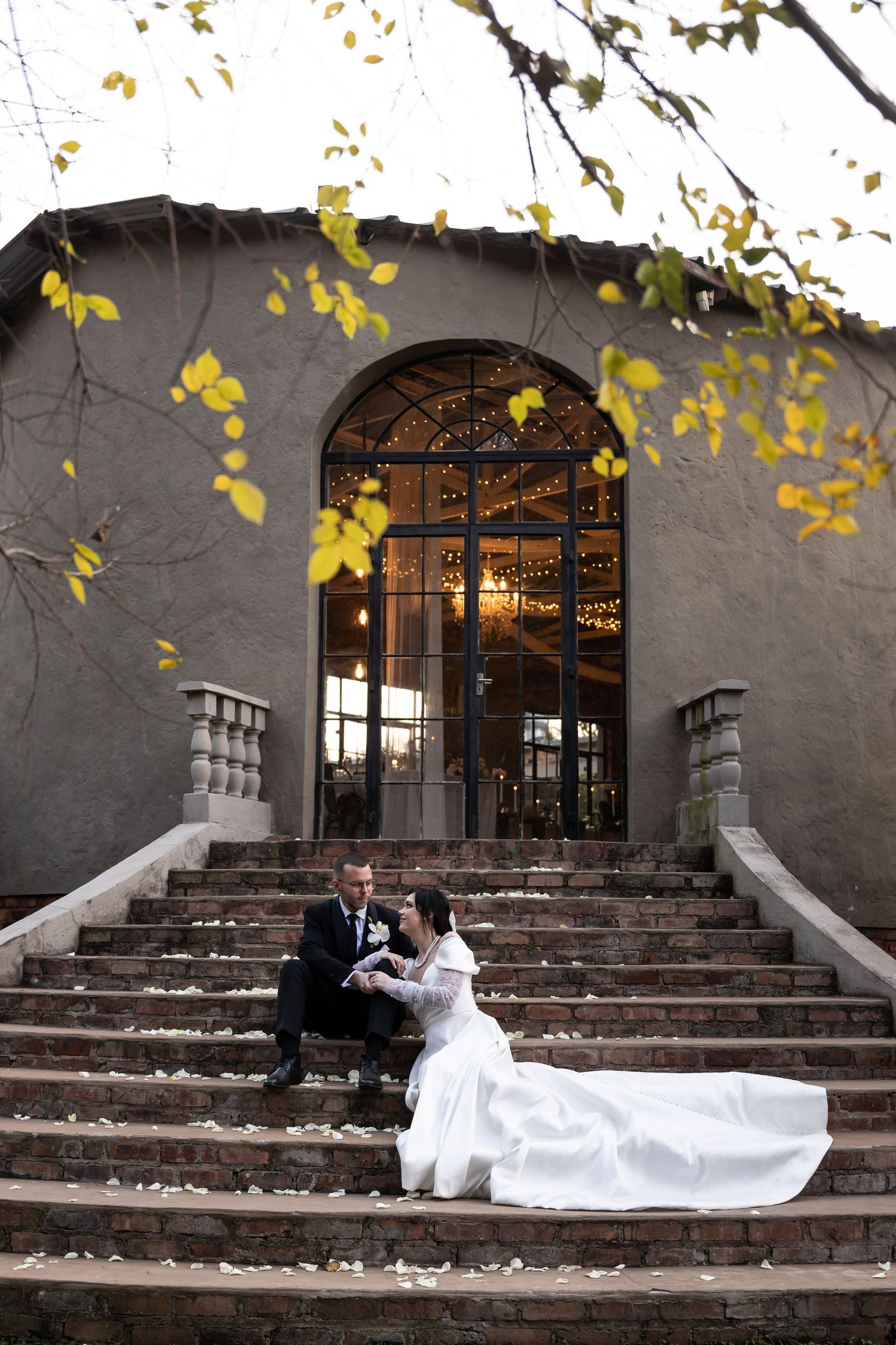 A couple sits on grand stone steps during an autumn wedding with yellow leaves and an arched entrance behind them.