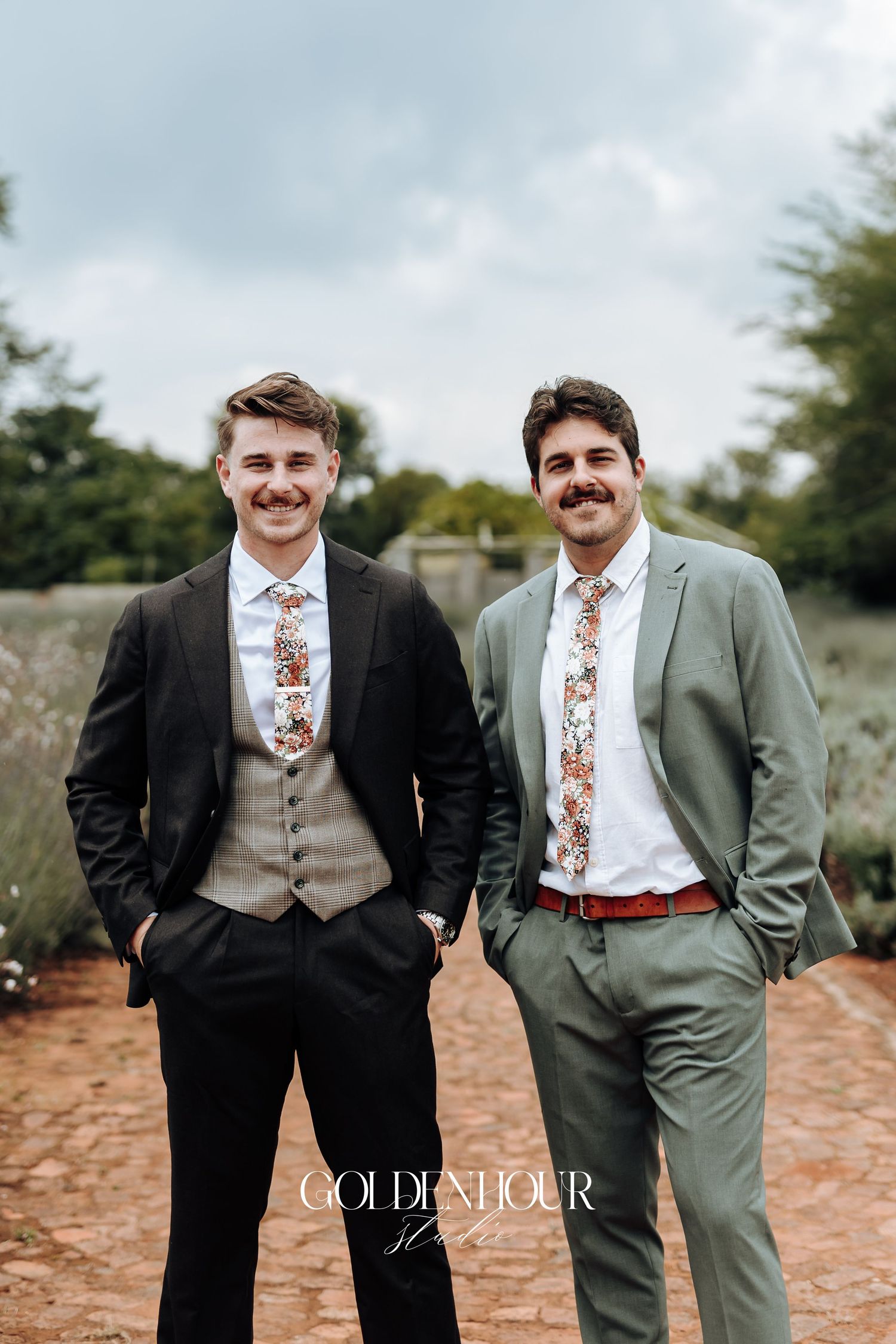 Two groomsmen in suits and floral ties pose together outdoors against a cloudy sky.