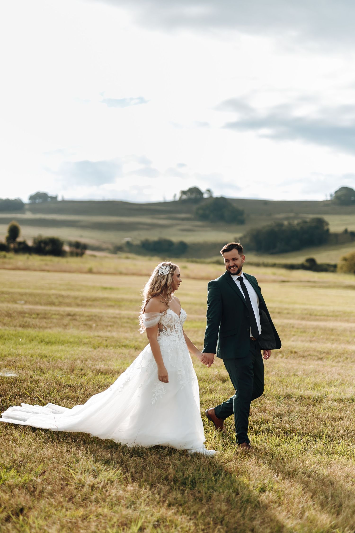 A bride and groom walk through a golden field at sunset with rolling hills in the background.
