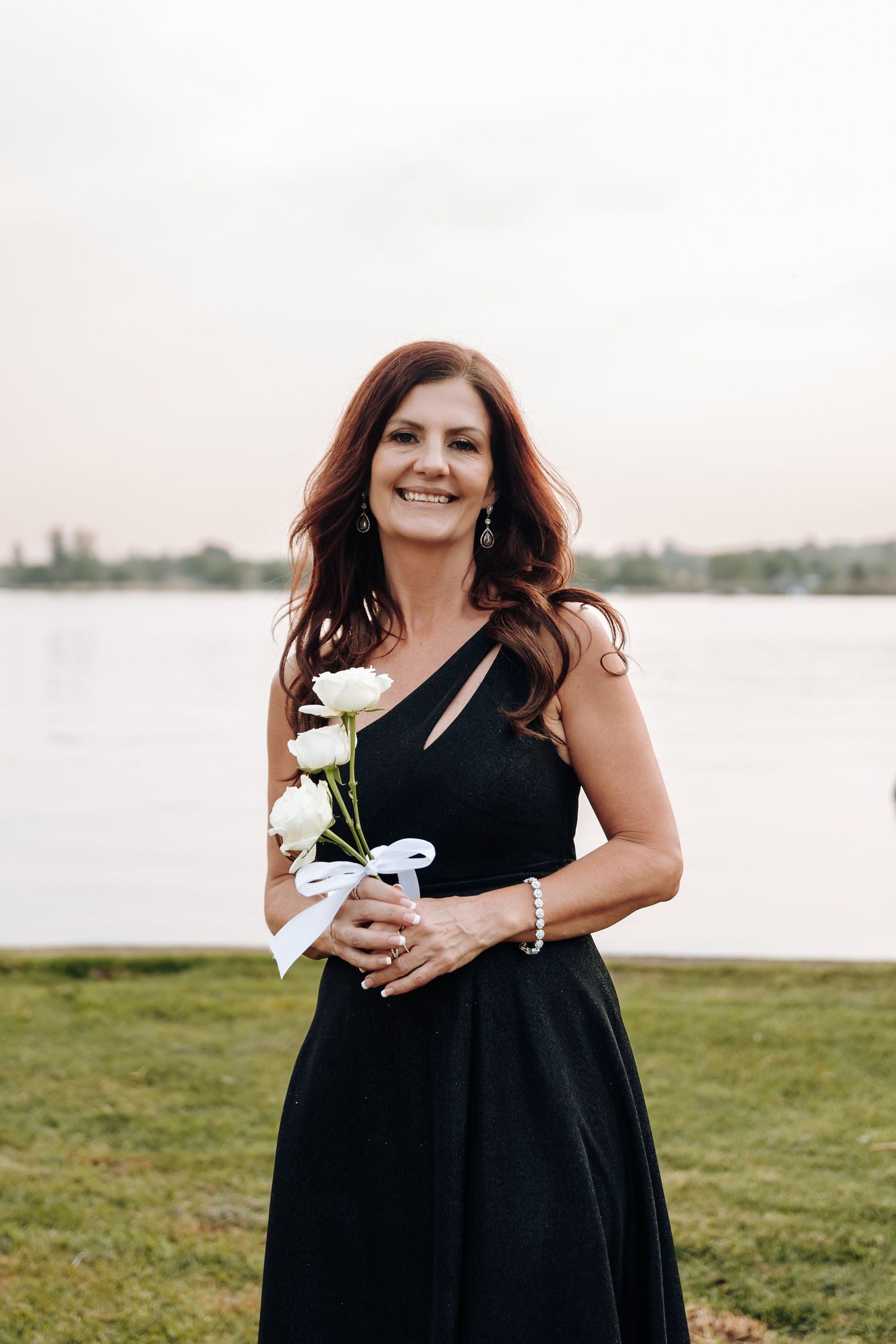 Woman in flowing black dress stands in grassy field by waterfront holding white flowers at sunset.
