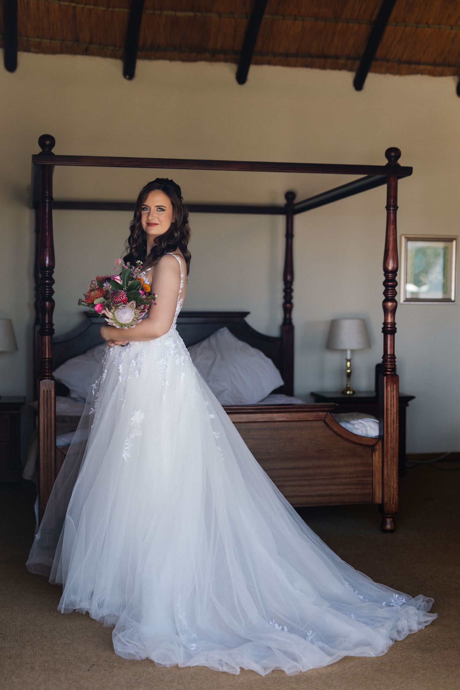 A bride in a flowing white tulle wedding gown poses in front of a wooden four-poster bed.